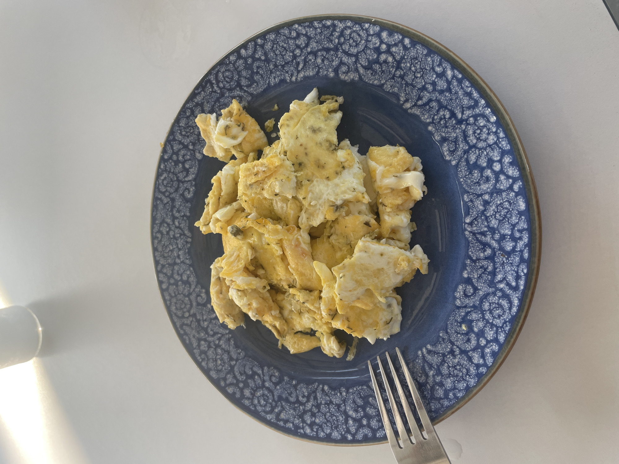 A plate of scrambled eggs is placed on a white table. The plate is dark blue with a white patterned rim. A fork with four tines rests on the left side of the plate, partially submerged in the eggs. The eggs are fluffy and golden yellow, with visible herbs mixed in. The background shows a white surface with a soft shadow cast by an object out of frame to the top left. The lighting suggests an indoor setting, possibly a kitchen or dining area. The context of Skala Avdiron, Greece, implies a meal served in a Greek locale.