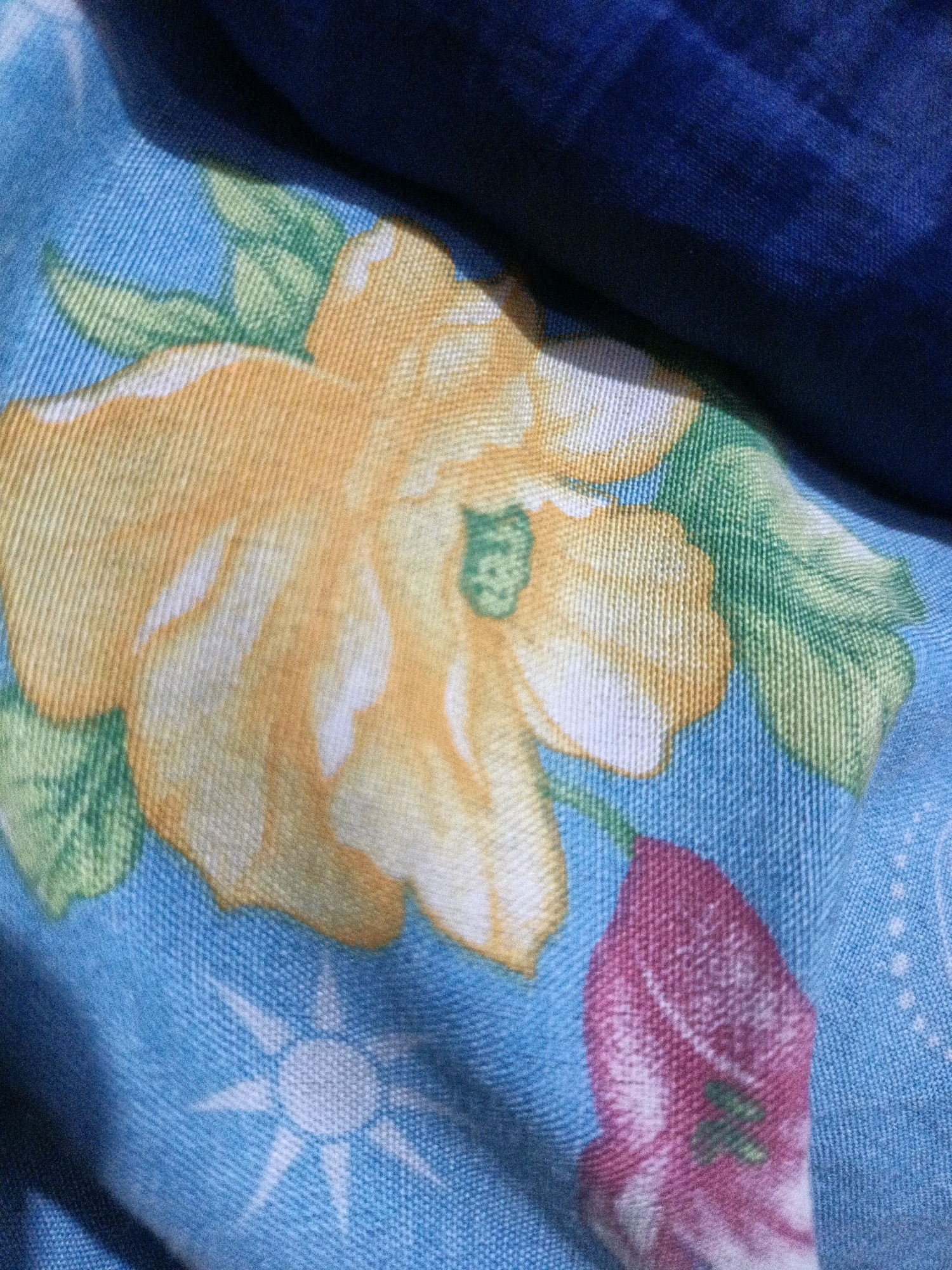 The image displays a close-up, high-angle shot of a floral patterned fabric. The fabric is predominantly blue, with a large, stylized yellow and white flower taking up a significant portion of the frame. There are also hints of green leaves and a pink petal visible. A dark blue fabric is draped over the top right portion of the patterned fabric. The texture of the fabric, with visible woven threads, is evident throughout the image. There are no people, actions, or interactions depicted, and the scene does not provide any information about a specific location in West Jakarta, Indonesia.