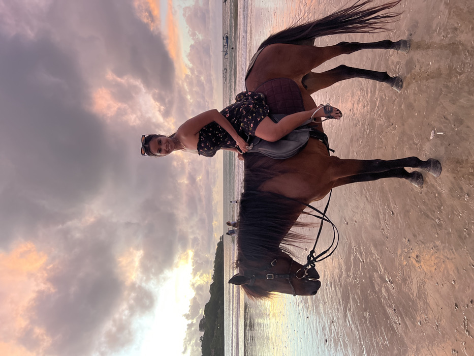 An East Asian female is seated on a bay horse on a wet sandy beach in Jimbaran, Indonesia, under an overcast sky displaying pink, orange, and purple hues indicative of sunset. The woman, appearing to be in her mid-20s to mid-30s, smiles directly at the camera. She wears a dark, strapless, floral-patterned mini-dress, a necklace, and dark sandals. Sunglasses are perched atop her head. She holds the horse's black reins with her left hand while seated on a grey English-style saddle over a dark red saddle pad. The horse stands still, facing the left, with a dark mane and tail, and dark hooves. In the mid-ground, the ocean features gentle waves, several indistinct human figures wading in the water, and multiple traditional Indonesian fishing boats (Jukungs) further out. A vegetated landmass with a visible structure on its crest is present on the distant left horizon.
