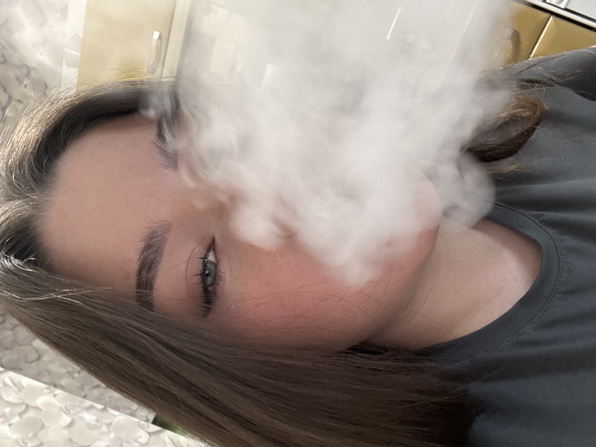A close-up, angled, indoor shot features a person with fair skin, light brown hair, and green eyes partially obscured by a cloud of white smoke. The person is wearing a dark gray shirt. The background appears to include light-colored walls. No information about the city of Perm, Russia is present in the image.