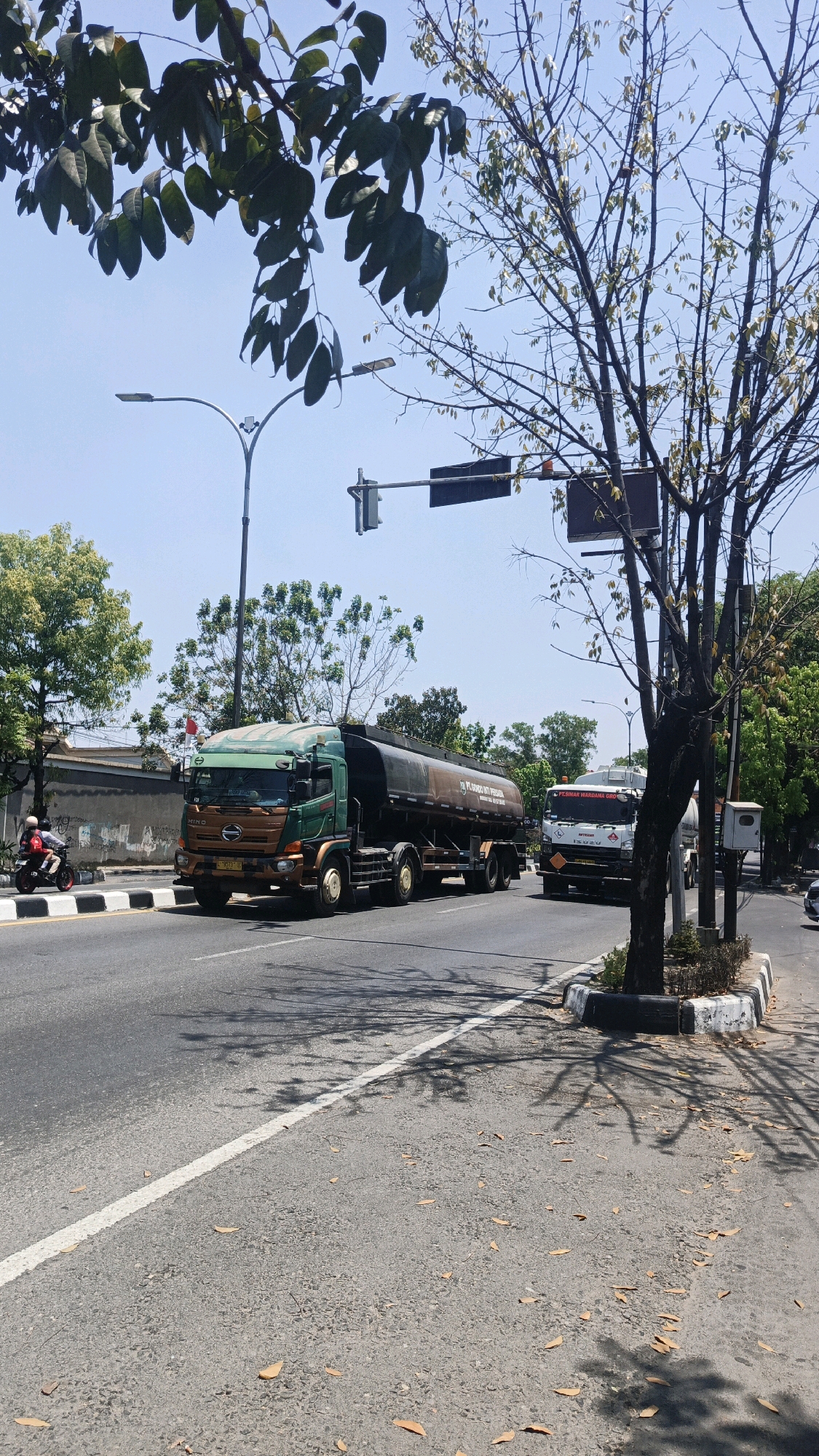 A Hino tanker truck, painted green and brown, drives on the right side of a paved road in Surakarta, Indonesia. The truck is carrying a dark-colored liquid in its cylindrical tank, which bears the inscription "PT. SUMBER MU PERTAMA". On the left side of the road, a motorcyclist is visible. Traffic lights are present overhead, and the sky is clear and blue. Trees line the right side of the road, and their shadows are cast on the asphalt. The scene appears to be a daytime view of a street in a city.