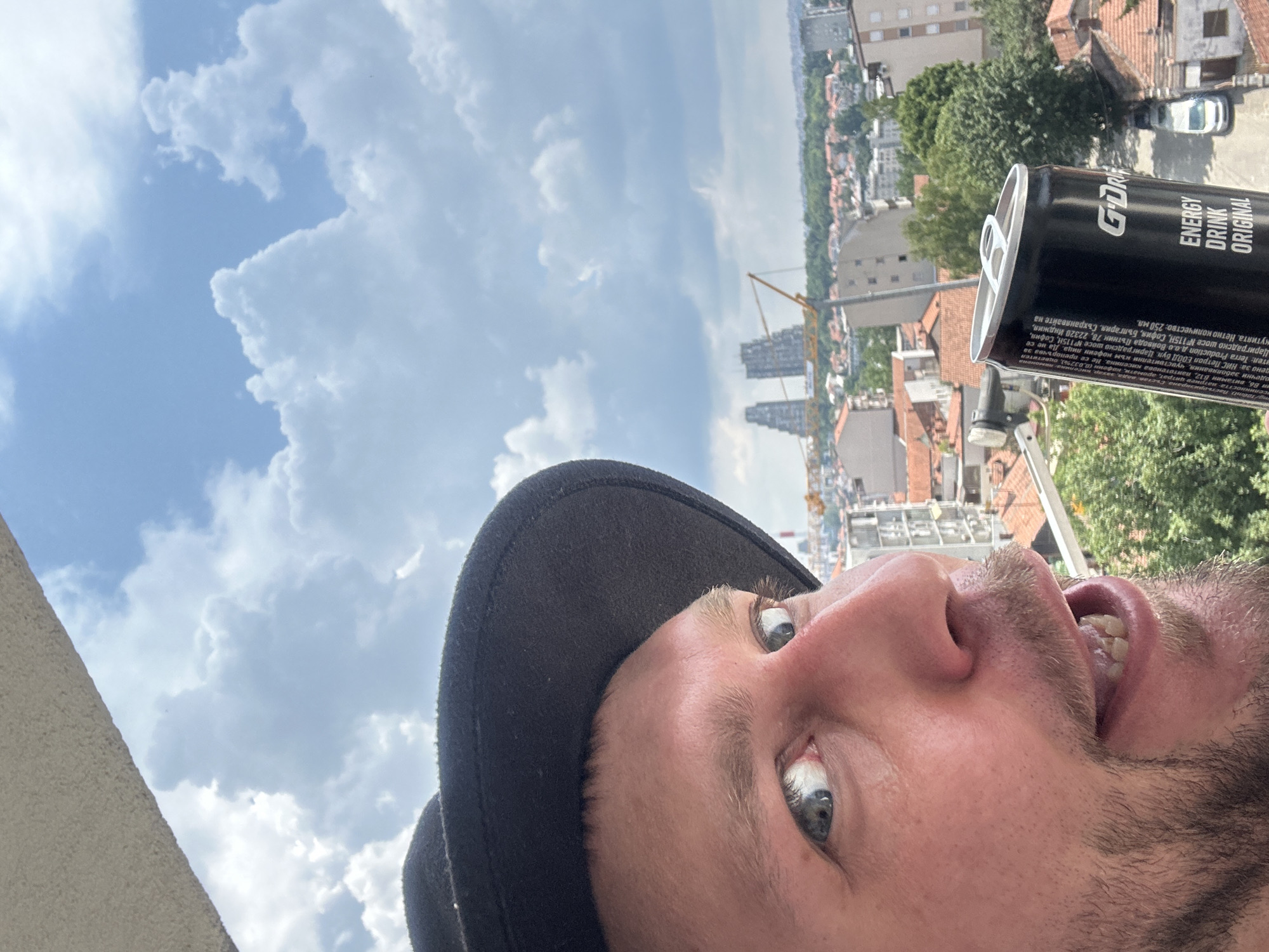 A man wearing a black hat and holding a can of G-DRIVE energy drink is looking off to the side against a backdrop of a city skyline under a cloudy sky. The city appears to be located in the Zvezdara Urban Municipality of Serbia, with buildings and trees visible in the distance. The man's face is partially visible, with his eyes and mouth open. The energy drink can is black with white text. The overall impression is a casual, outdoor scene in an urban environment.