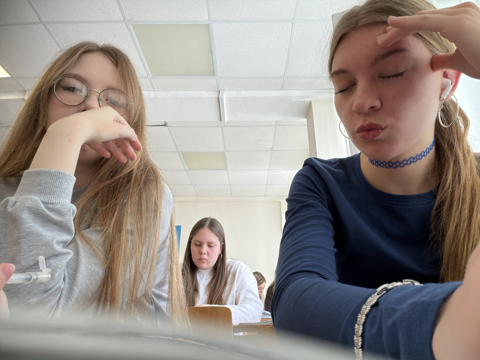 The image depicts a classroom scene with three young females visible. In the left foreground, a female with long, light brown hair and round eyeglasses wears a grey long-sleeved top. Her left hand is raised, partially covering her chin and mouth, while her right hand holds a transparent, cylindrical object, possibly a vape pen, near her shoulder. To her right, in the immediate foreground, a second female with long brown hair wears a dark blue long-sleeved top, a blue patterned choker necklace, and silver hoop earrings. She has a white earbud in her right ear. Her eyes are closed, and her lips are pursed in a pouting expression, while her right hand rests against her forehead/temple area. A silver-colored bracelet with decorative links is visible on her left wrist. In the midground, centered between the two foreground subjects, a third female with long dark hair, wearing a white long-sleeved top and silver hoop earrings, is seated at a light brown wooden desk, looking downwards, seemingly focused on materials on the desk. Additional blurred figures are visible in the background, further indicating a group setting. The ceiling features white acoustic tiles with recessed rectangular fluorescent light fixtures. The walls are plain white. The setting is consistent with a typical classroom or educational environment.