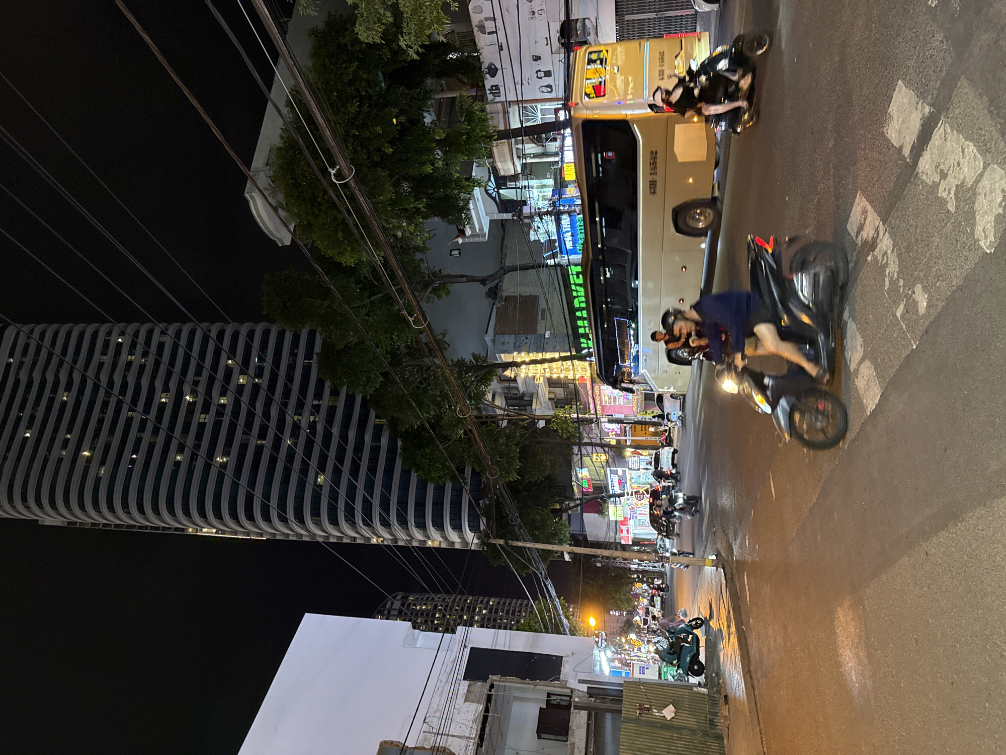 In the city of Nha Trang, Vietnam, at night, a busy street scene unfolds. A large, modern high-rise building with a distinctive curved facade dominates the background, its many windows illuminated. The street is illuminated by streetlights, neon signs from shops lining the road, and the headlights of vehicles. Multiple motorbikes, common in Vietnam, are the primary mode of transport. One motorbike is in the foreground, with a rider wearing a blue shirt and dark shorts, their legs visible. Another motorbike is in the middle ground, with a rider wearing a black helmet and dark clothing. A light-colored tour bus, marked with Vietnamese license plates, is moving down the street. Several other motorbikes and cars are also visible, suggesting a lively urban environment. The road surface appears wet, reflecting the streetlights and vehicle lights, possibly from recent rain. Trees with dense foliage are interspersed along the street. The overall impression is of a vibrant and active urban nightscape.