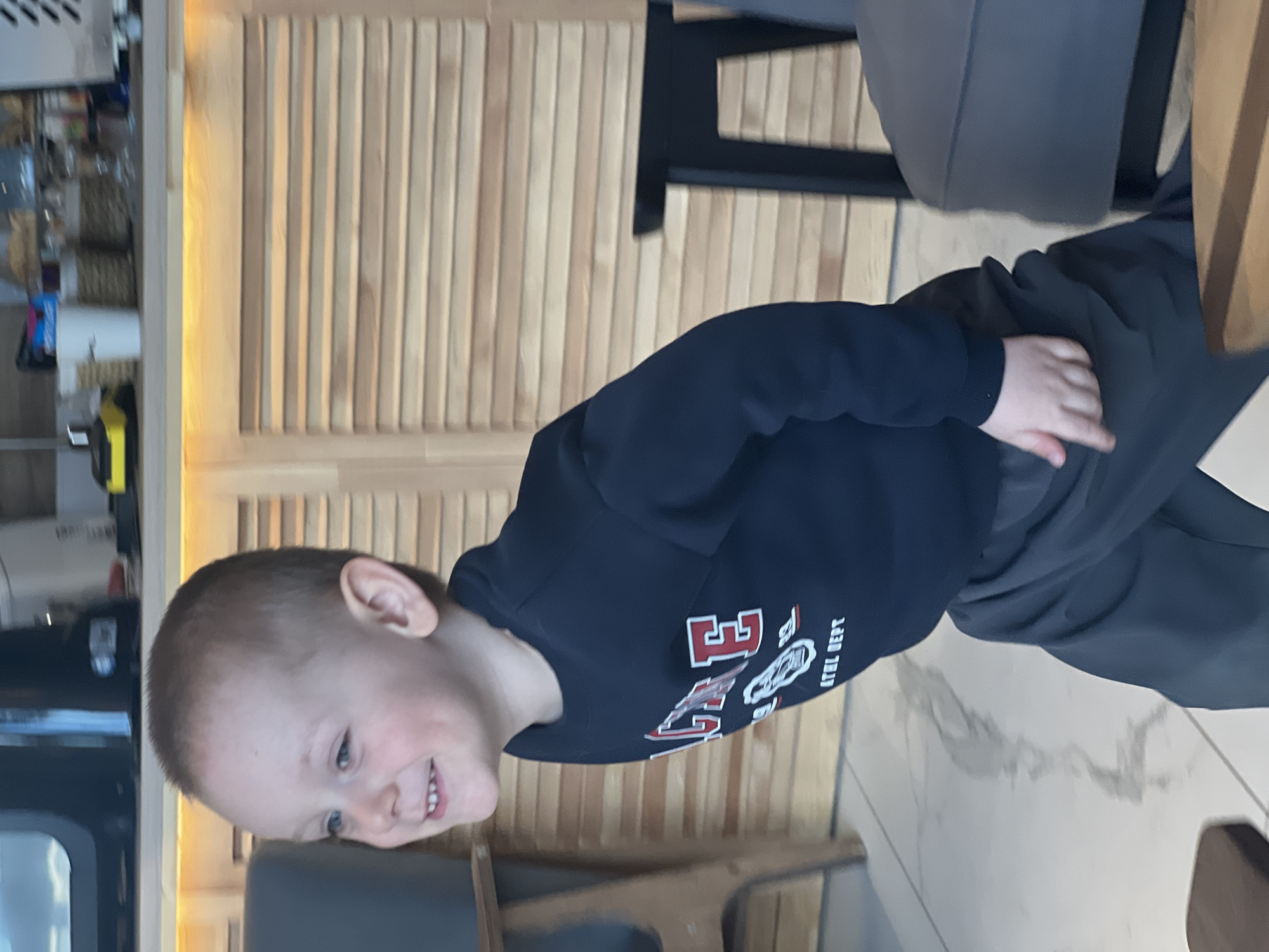 A young boy is standing in a room in Mytishchi, Russia. The room has a wooden wall and a light-colored tile floor. The boy is wearing a dark sweatshirt with a red and white logo. He is smiling and looking to the left. A kitchen or bar area is partially visible in the background. Furniture, including chairs and a table, is present.