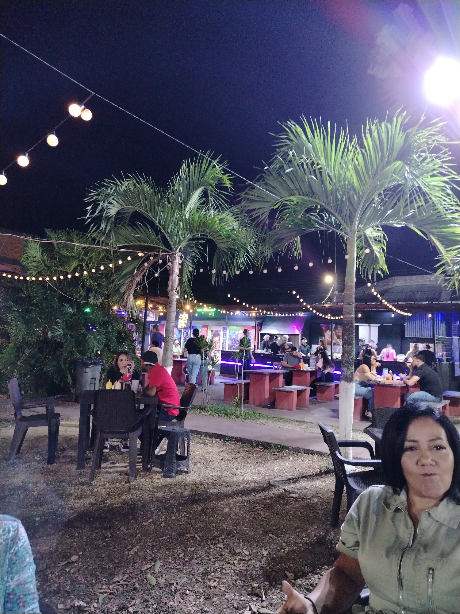 The image depicts an outdoor gathering at night, possibly in the city of Tinaquillo, Venezuela. The scene includes several individuals seated at tables, suggesting a social setting like a restaurant or bar. String lights illuminate the area, creating a festive atmosphere. Palm trees are visible, indicating a tropical or subtropical location. People are interacting, some are eating or drinking, while others are engaged in conversation. The ground appears to be gravel or dirt. A woman is seated in the foreground, seemingly part of the gathering. In the background, there's a building or structure with activity, potentially a bar or food service area.