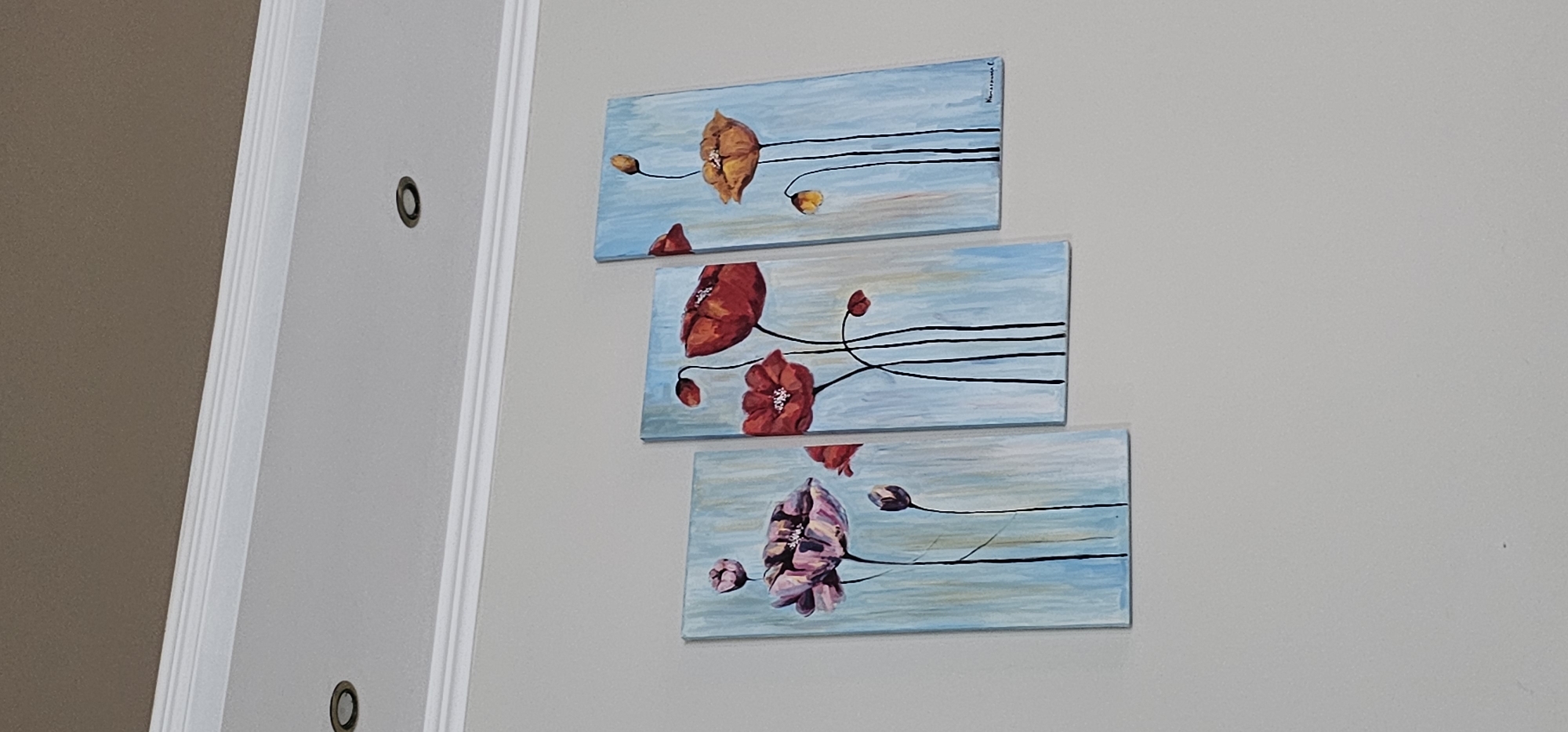 The image shows a triptych painting of poppies hanging on a light gray wall. The painting is composed of three vertical panels, each depicting stylized poppies against a light blue background with vertical brushstrokes. The leftmost panel features a large, open pink poppy and a closed bud. The middle panel displays two large, vibrant red poppies, one open and one slightly unfurled, along with a smaller bud. The rightmost panel shows a large, open orange-yellow poppy and two smaller buds, one yellow and one orange. Thin black lines represent the stems of the poppies. The top of the image shows a white crown molding and a portion of a brown ceiling with two recessed light fixtures. The overall impression is one of modern floral art displayed in an indoor setting.