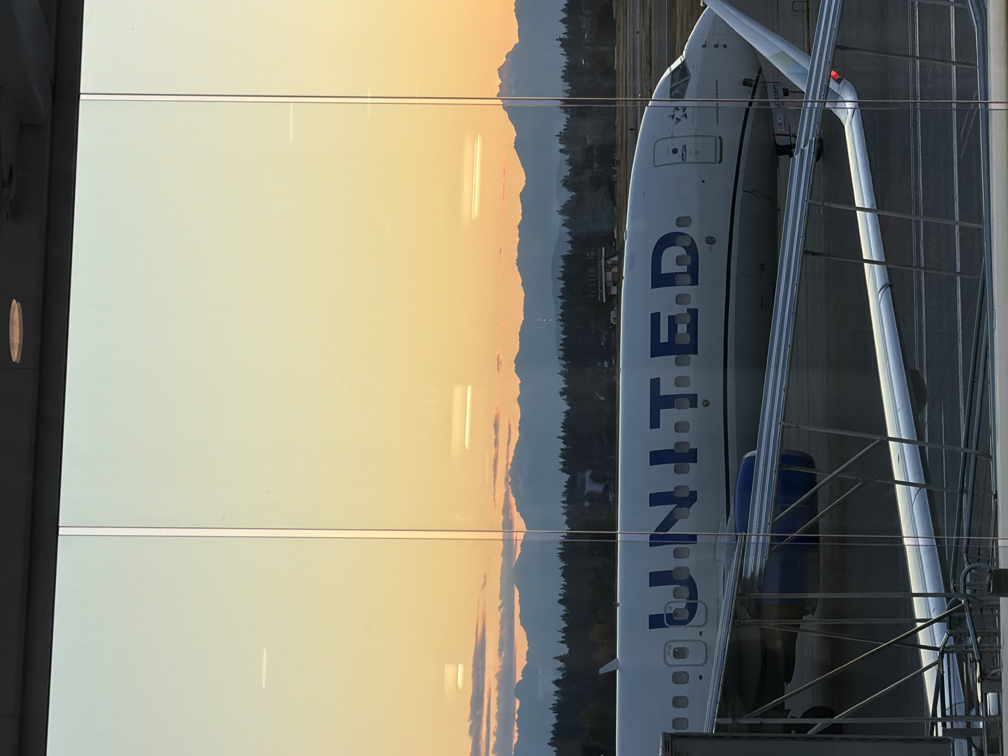 The image shows a United Airlines airplane parked at Sea-Tac International Airport in Seattle, Washington, USA. The plane's fuselage, with the "UNITED" logo clearly visible, is positioned in the foreground. Part of the wing and an engine are also visible. In the background, a scenic view of mountains and trees is seen against a golden-orange sunset sky, with clouds scattered across the horizon. A jet bridge extends towards the aircraft, and a portion of the airport terminal's glass facade is visible, reflecting the sky and the surrounding environment. The scene appears to be captured from inside the terminal, looking out at the tarmac.