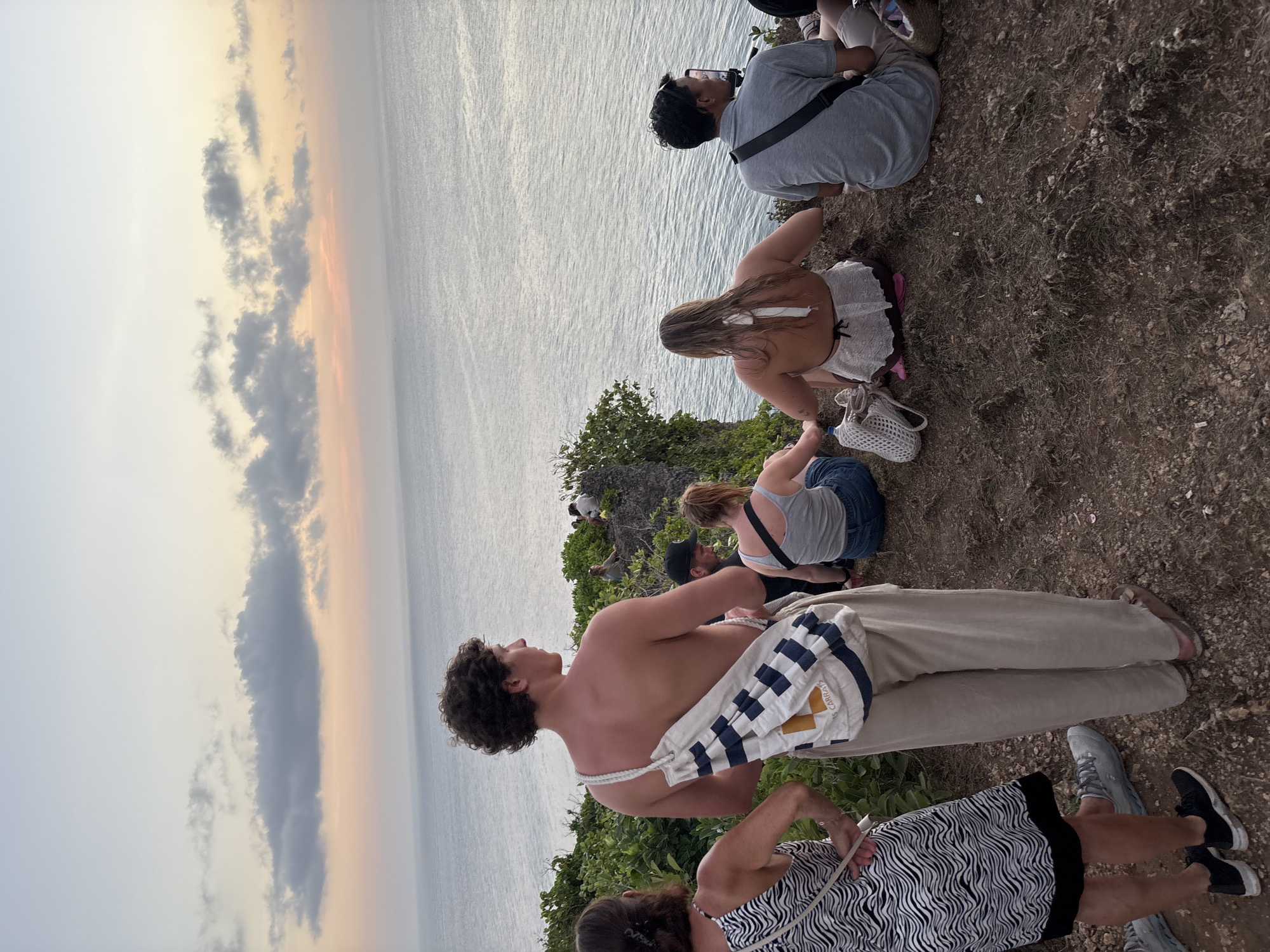 An evening scene depicts a group of individuals sitting on a cliff overlooking the ocean near Kuta Selatan, Indonesia. The sky displays a gradient of pastel colors, transitioning from a pale blue at the top to hues of pink and orange near the horizon. Several people are seated on the grassy, rocky terrain. In the foreground, a shirtless man with curly hair stands facing the ocean, carrying a white and blue striped bag over his shoulder. To his left, a woman in a black and white patterned dress stands facing away from the camera. Other individuals are seated in the middle ground, some facing the ocean and others looking towards the side. The ocean stretches out to the horizon, with gentle ripples visible on its surface. The overall atmosphere suggests a moment of shared observation of the sunset.