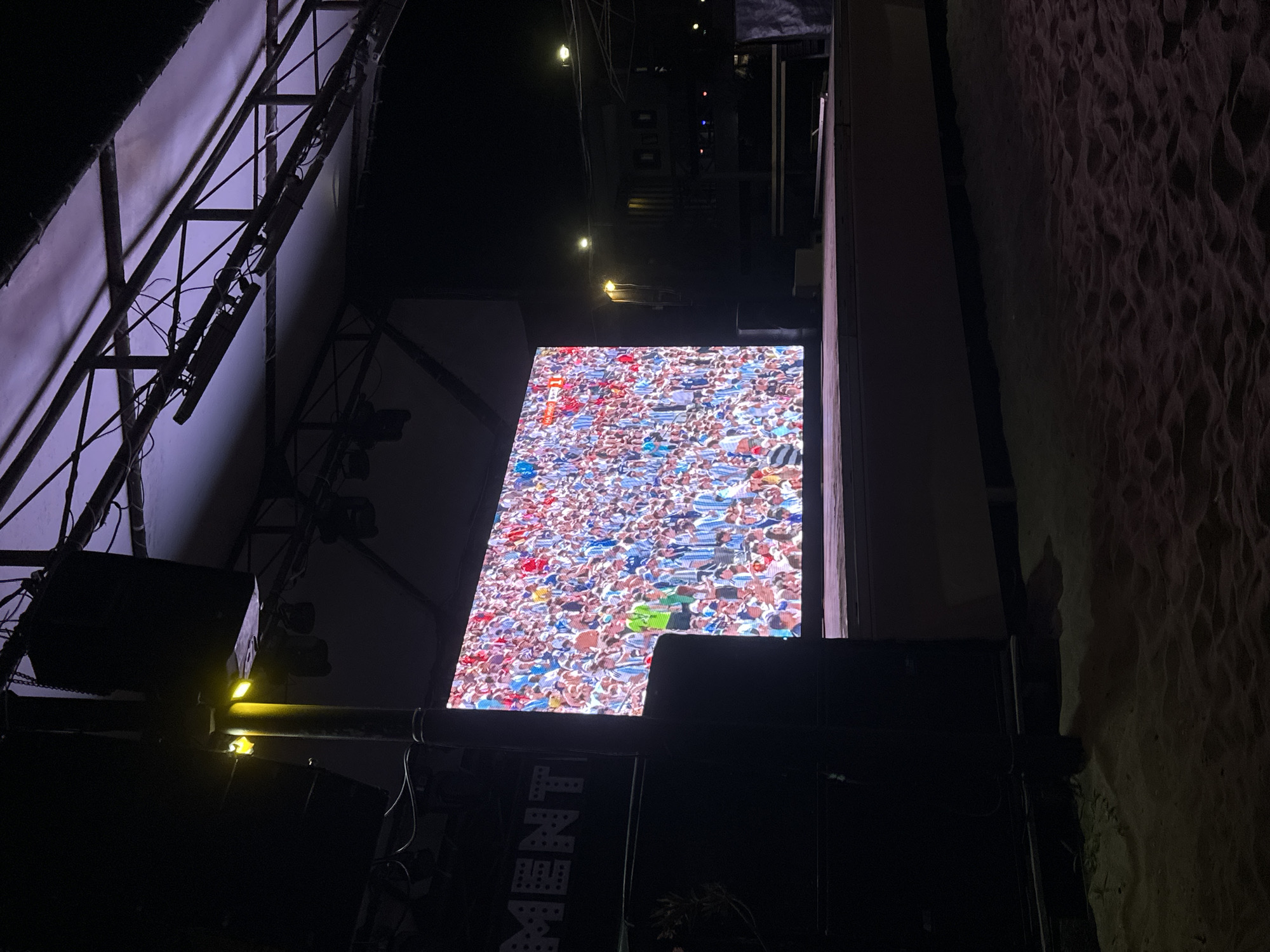 The image captures an outdoor stage at night, likely on a beach or sandy area, given the texture of the ground in the foreground. A large LED screen dominates the stage, displaying a densely packed crowd of people, many wearing light blue and white striped shirts, suggesting a sporting event or a fan gathering. The screen is illuminated, casting a glow onto the stage platform. Above the stage, a metal truss structure supports speakers and lighting equipment, partially covered by a translucent material that appears to be lit from above with a purple hue. To the left of the stage, a black structure with white lettering that reads "MENT" is visible, possibly part of a larger banner or signage. The overall scene suggests an event setup for a public viewing or performance in an open-air venue in Varna, Bulgaria.
