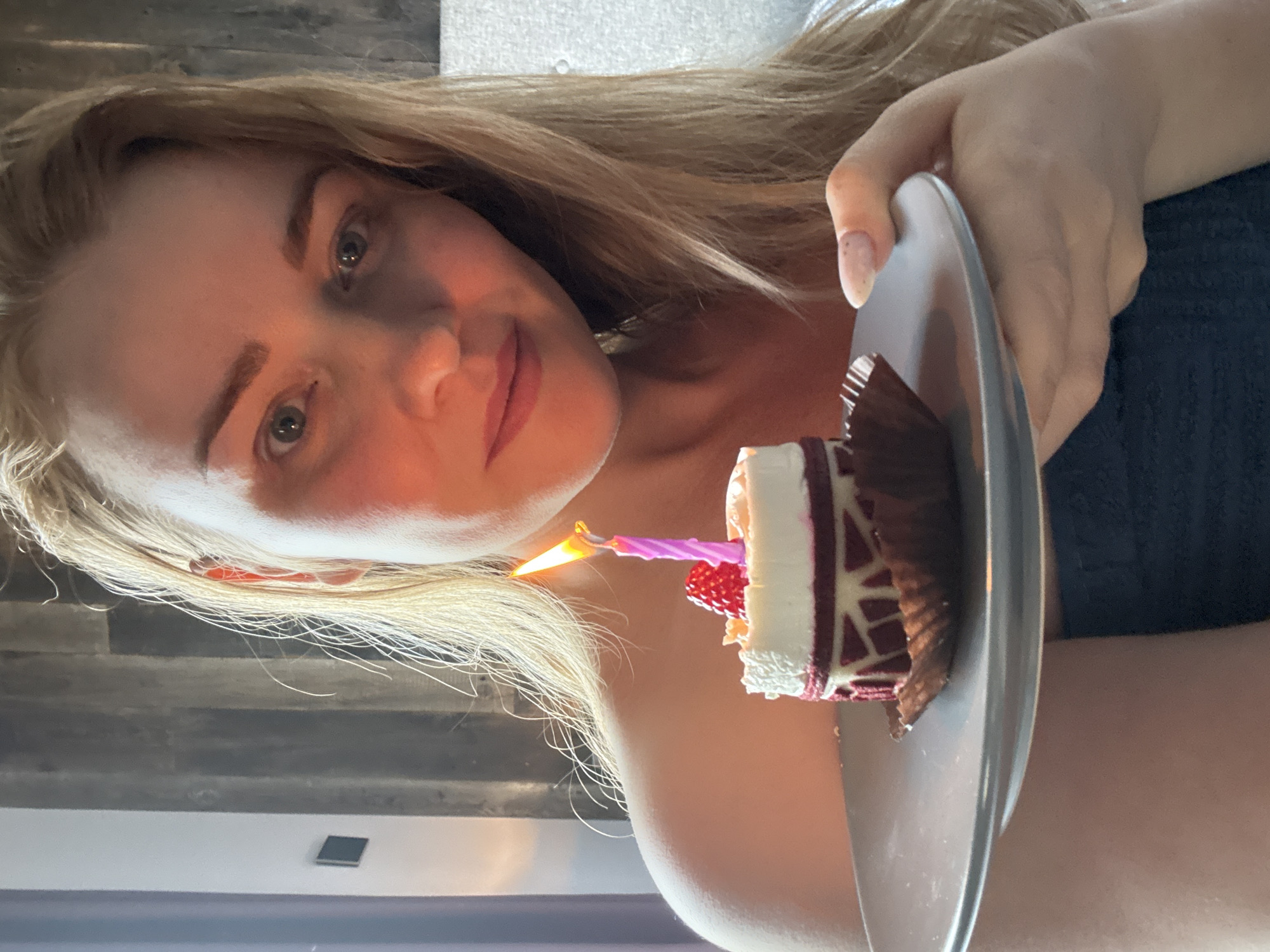 A fair-skinned person with light brown hair is holding a small piece of cake with a lit candle on a gray plate. The cake appears to be a multilayered dessert with white frosting and dark chocolate. The person is smiling and looking directly at the camera. The background includes a wooden-like wall and a lighter-colored surface, possibly a wall or curtain. There is not enough information to determine if the image was taken in Saint Petersburg, Russia.