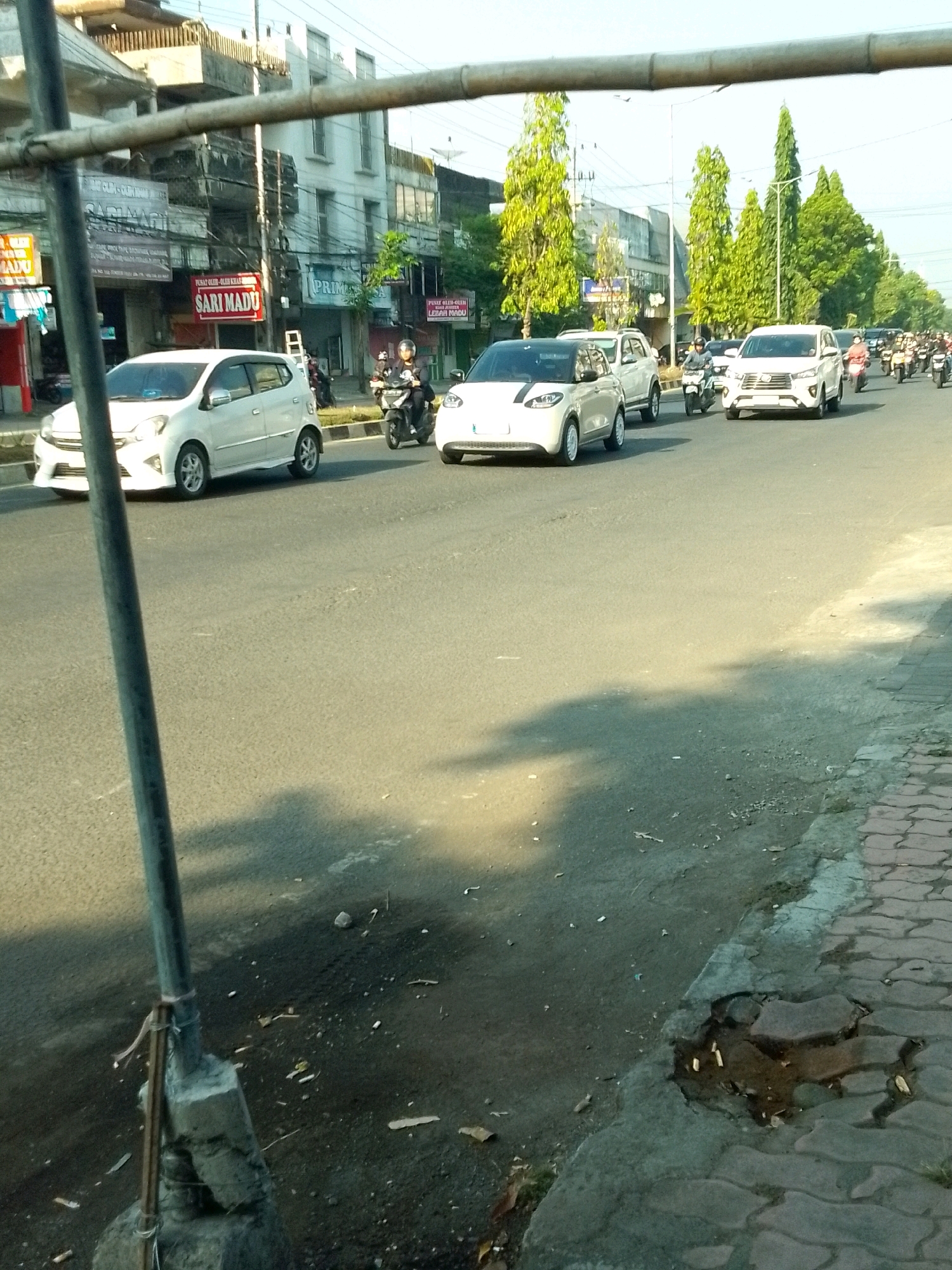 A street scene in Jember, Indonesia, shows traffic moving along a paved road lined with buildings and trees. On the left, a white car is parked. In the middle of the road, a white small car is driving, followed by a white SUV and several motorcycles. The road is bordered by a sidewalk on the right, which is partially cracked and has some debris. The buildings on the left appear to be commercial establishments with signs, including one that says "SARI MADU". Overhead, a metal structure, possibly part of a temporary fence or scaffolding, runs across the top of the image. The sky is clear and bright, suggesting it is daytime.