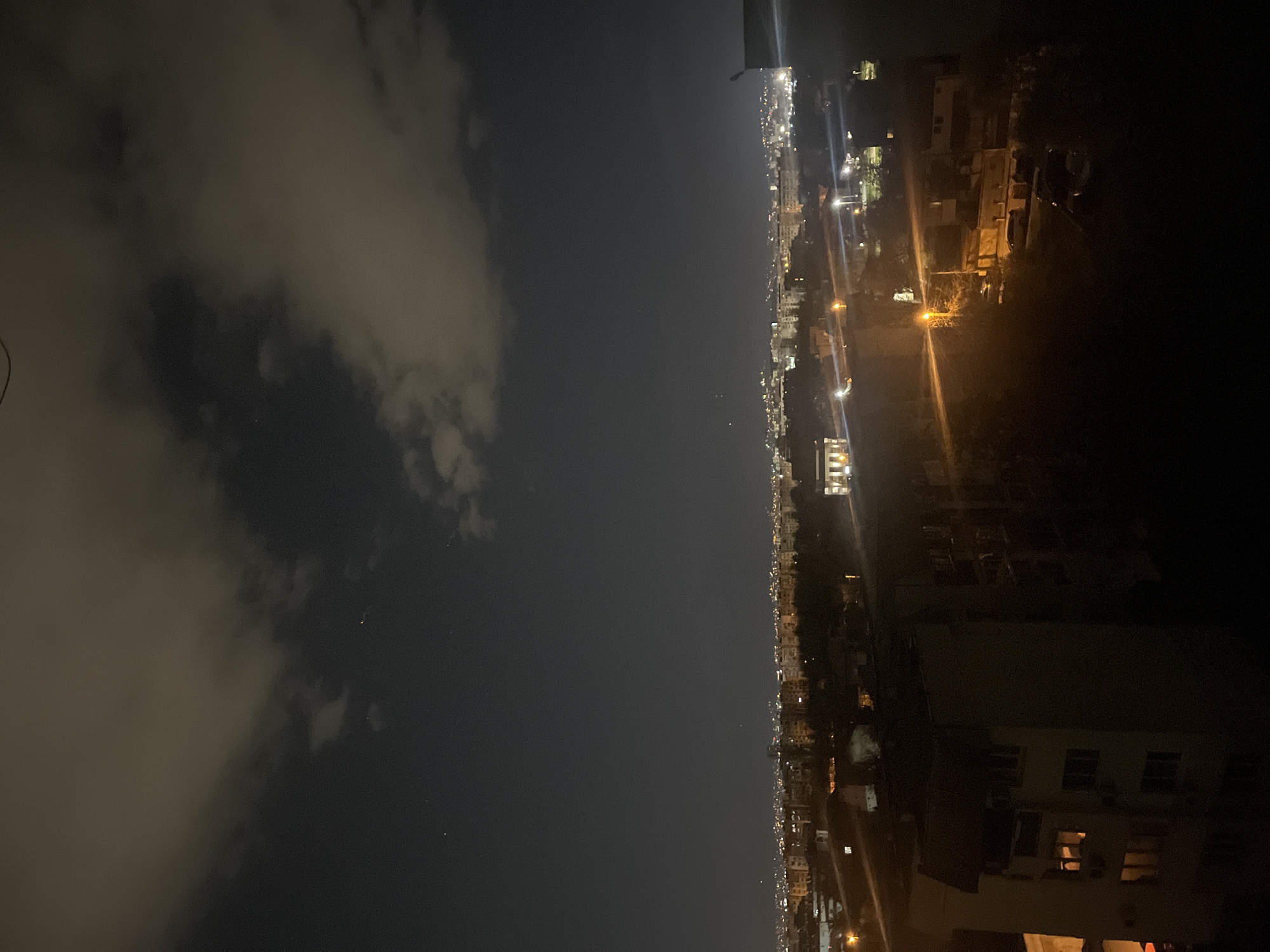 Here's a description of the image:
The image is a nighttime view from a high vantage point, likely a rooftop or high-rise balcony, showing a city skyline and sky. The city is illuminated with streetlights and lights from buildings, stretching into the distance. The right portion of the image is dominated by a building close to the vantage point, with illuminated windows. The sky is dark with some cloud cover on the left portion of the image. It is not possible to determine the specific location in Sofia, Bulgaria with only the provided information.
