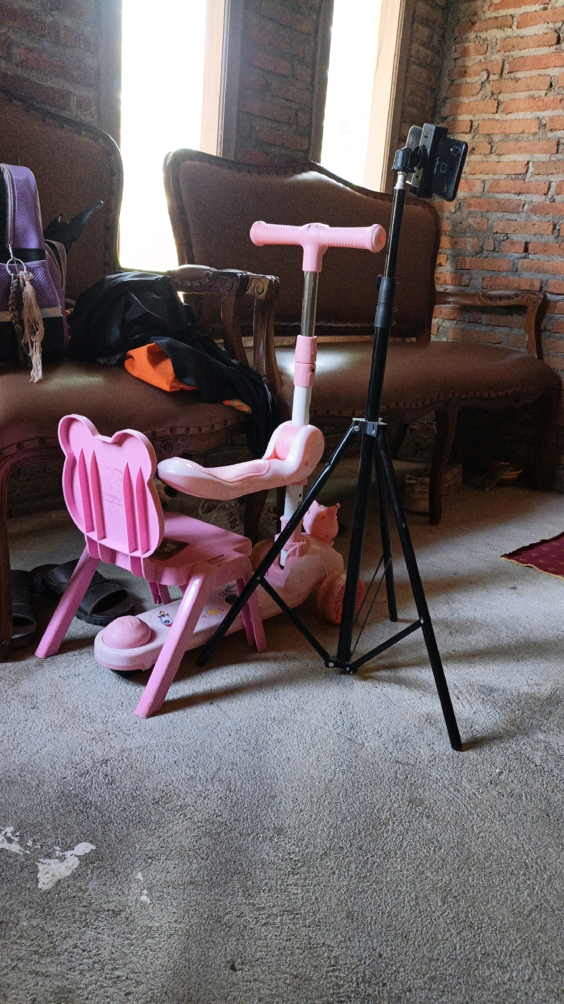 The image depicts an indoor scene, likely a room in a residence or studio, with exposed brick walls and a concrete floor. The primary subjects are a pink children's scooter with a detachable seat and backrest, and a black tripod with a mobile phone mounted on top. The scooter is positioned in the foreground, facing left, and appears to be a convertible model that can be used as a ride-on toy or a push scooter. Behind the scooter, two upholstered chairs, possibly antique, are visible, one with a purple backpack draped over its arm. The tripod stands to the right of the scooter, angled slightly towards the right, suggesting it's set up for recording or photography. The overall lighting suggests it's daytime, with light filtering in from an unseen window. There are no people in the frame. The location is identified as Cikal, Indonesia, which suggests a residential or commercial setting within that city.
