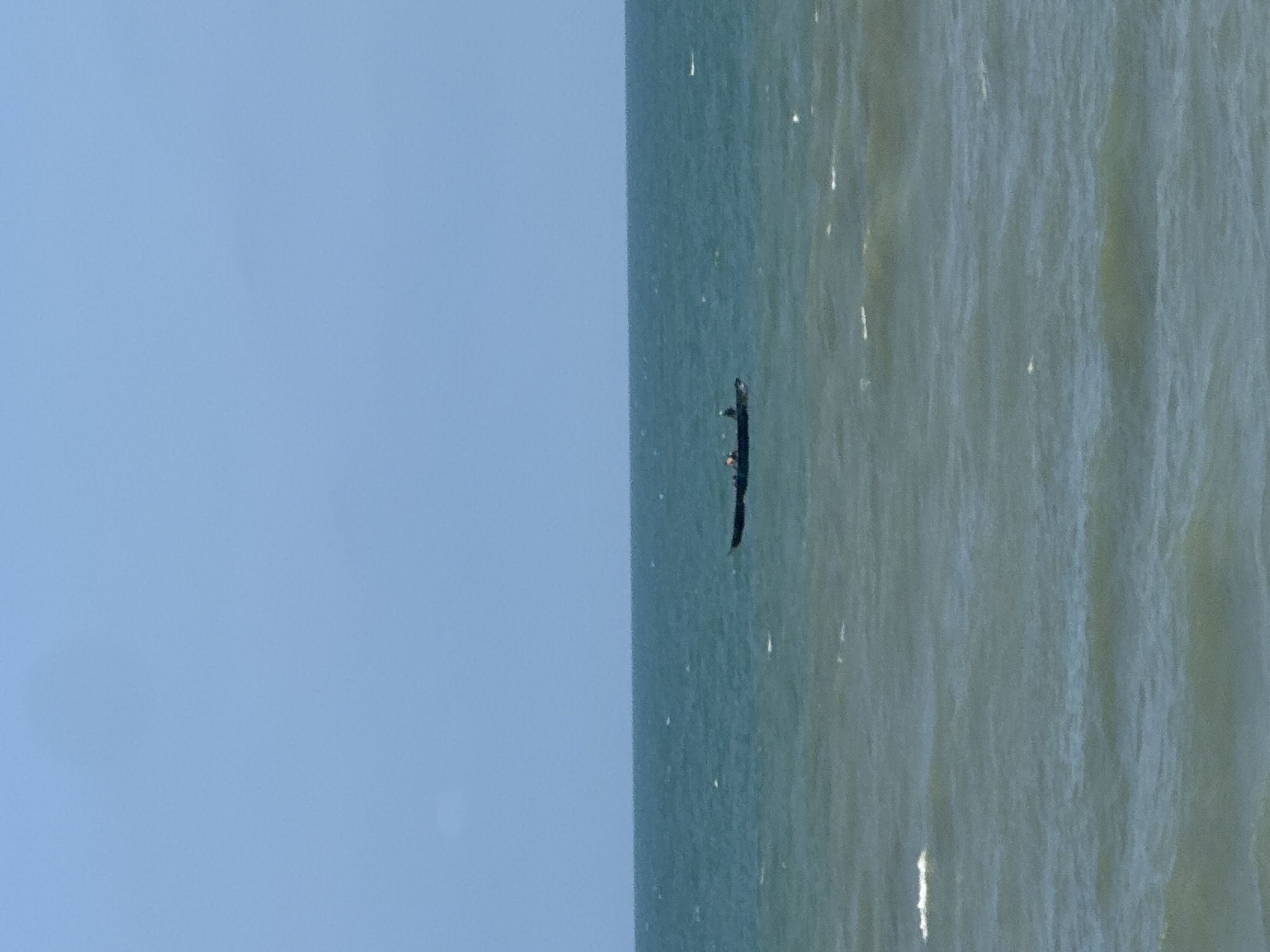 The image shows a person in a canoe on the water. The canoe is dark and appears to be moving. The water is a deep blue-green color, with gentle waves reflecting the sunlight. The sky is a clear, light blue. The scene is serene and peaceful, with the vast expanse of water stretching out to the horizon. It is difficult to determine the exact time of day or weather conditions, but the presence of sunlight suggests it is daytime with fair weather. The location is identified as Badagry, Nigeria, which implies the setting is likely a coastal or inland waterway. The image captures a solitary moment of transit on the water, with the focus on the person and their mode of transport. There is no visible text in the image.