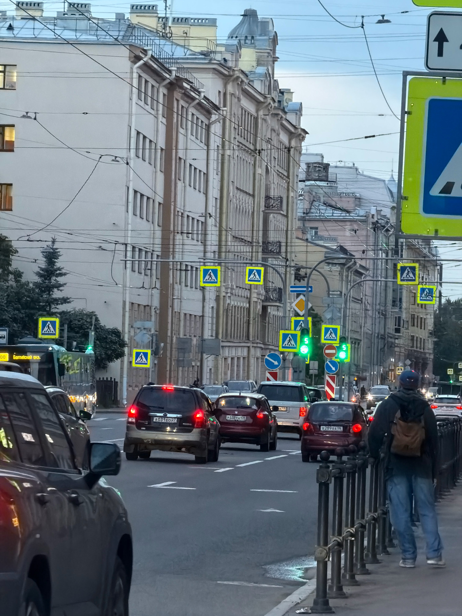 The image depicts a street scene in Saint Petersburg, Russia, likely during dusk or overcast weather. The perspective is from a street level, looking down a wide avenue lined with multi-story buildings, characteristic of Saint Petersburg's architecture with classical facades and mansard roofs. Traffic is moderate, with several cars and a bus visible on the road. Pedestrian crossing signs, marked with yellow squares and white figures walking, are prominently displayed at intervals, indicating crosswalks. Traffic lights, some showing green, are also present. A man with a backpack is walking on the sidewalk, leaning on a decorative metal fence. The overall impression is a typical urban environment with functional infrastructure and a sense of everyday activity.