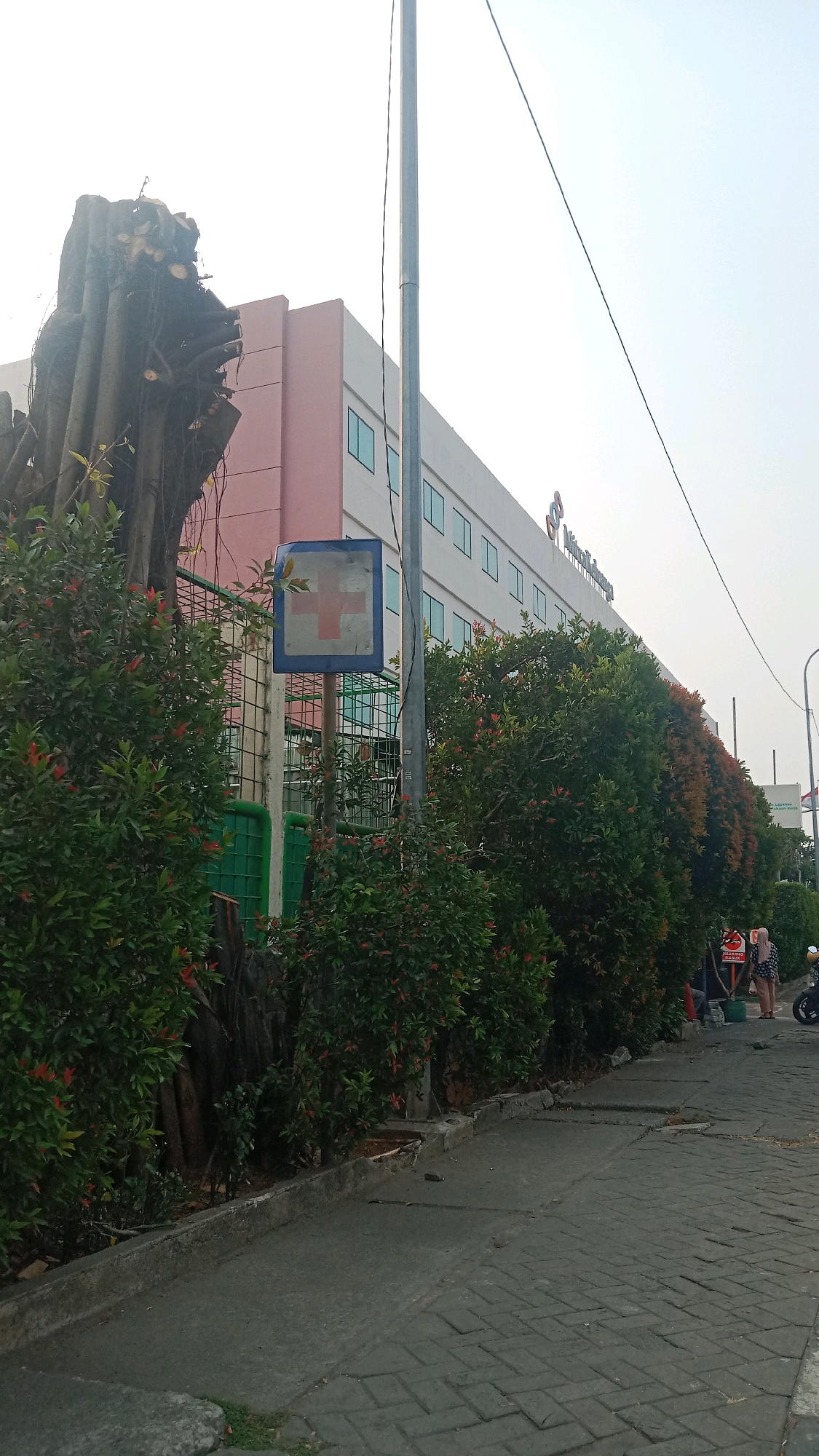 The image depicts a street scene in Bekasi, Indonesia, with a building on the right and lush greenery on the left. The building appears to be a medical facility, as indicated by a prominent red cross sign on a blue square plaque mounted on a pole. The building itself is a multi-story structure with a light pink and white facade, featuring rows of rectangular windows. The greenery consists of dense bushes and trees, some of which have small red flowers, lining the sidewalk and surrounding the base of the building. In the foreground, a paved walkway leads towards the building, with a concrete curb separating it from the vegetated area. A tall metal pole stands near the center of the image, with wires extending upwards. On the far right, two individuals are visible near a motorcycle, and a sign with Indonesian text is partially visible. The sky is clear and overcast.