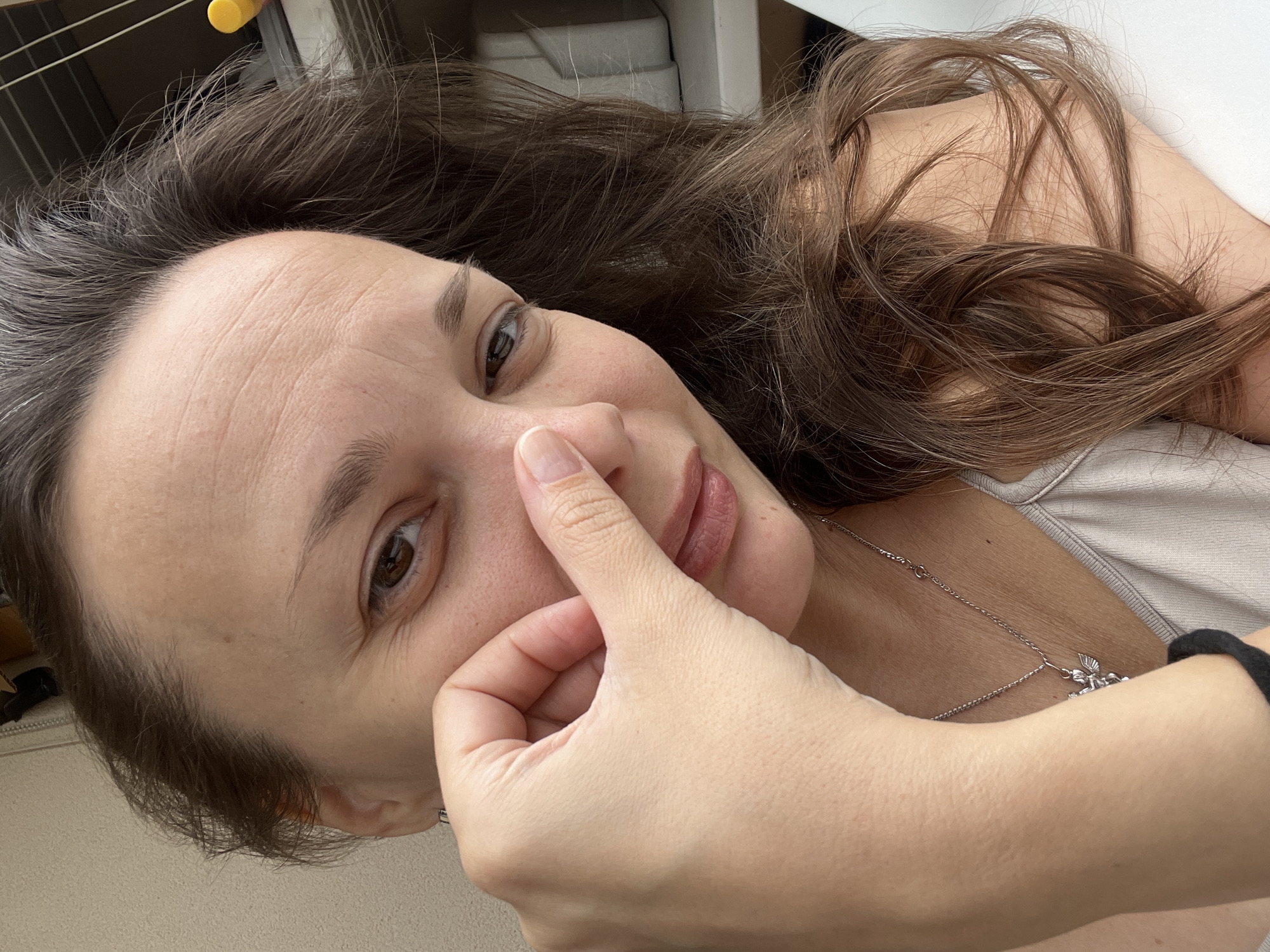 A close-up shot shows a woman with brown hair and brown eyes. Her right hand is near her face, with her thumb and index finger touching her nose. She is wearing a light-colored tank top with a silver necklace that has a small angel pendant. The background is blurry and appears to be indoors with some shelving and possibly a window. The lighting suggests it is daytime. There is no information in the image to suggest a location in Tolyatti, Russia.