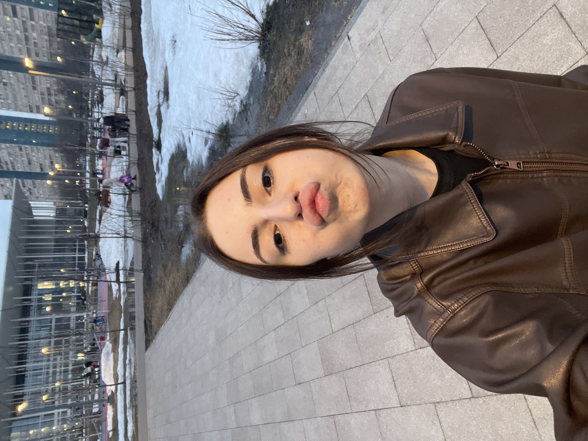 A young female subject is captured in a waist-up, slightly high-angle selfie, dominating the foreground. She has dark brown hair and is wearing a dark brown, zippered, possibly leather jacket over a black top. Her facial expression is characterized by pursed lips and slightly widened eyes.
The scene is set in an urban park, identified as Zaryadye Park in Moscow, Russia, during late winter or early spring at dusk. The ground consists of light-colored, rectangular paved pathways, with adjacent areas of bare earth, dry vegetation, and scattered patches of melting snow and ice. Numerous bare deciduous trees and bushes are visible throughout the mid-ground.
In the mid-ground, a modern building with a glass facade and distinctive vertical architectural elements, possibly a cultural or concert hall, stands prominently. Internal or external lights from this building are illuminated. To its right, a children's playground area with colorful equipment (red, pink, yellow) is visible. Several indistinct figures are present in the mid-ground: one person in a light-colored coat is seen walking, carrying a bag, while other figures are gathered near the playground equipment.
In the far background, several tall, multi-story residential buildings, characteristic of Soviet-era or modern urban development, rise against the twilight sky. Artificial streetlights or park lights are also visible in the background, adding to the evening illumination.