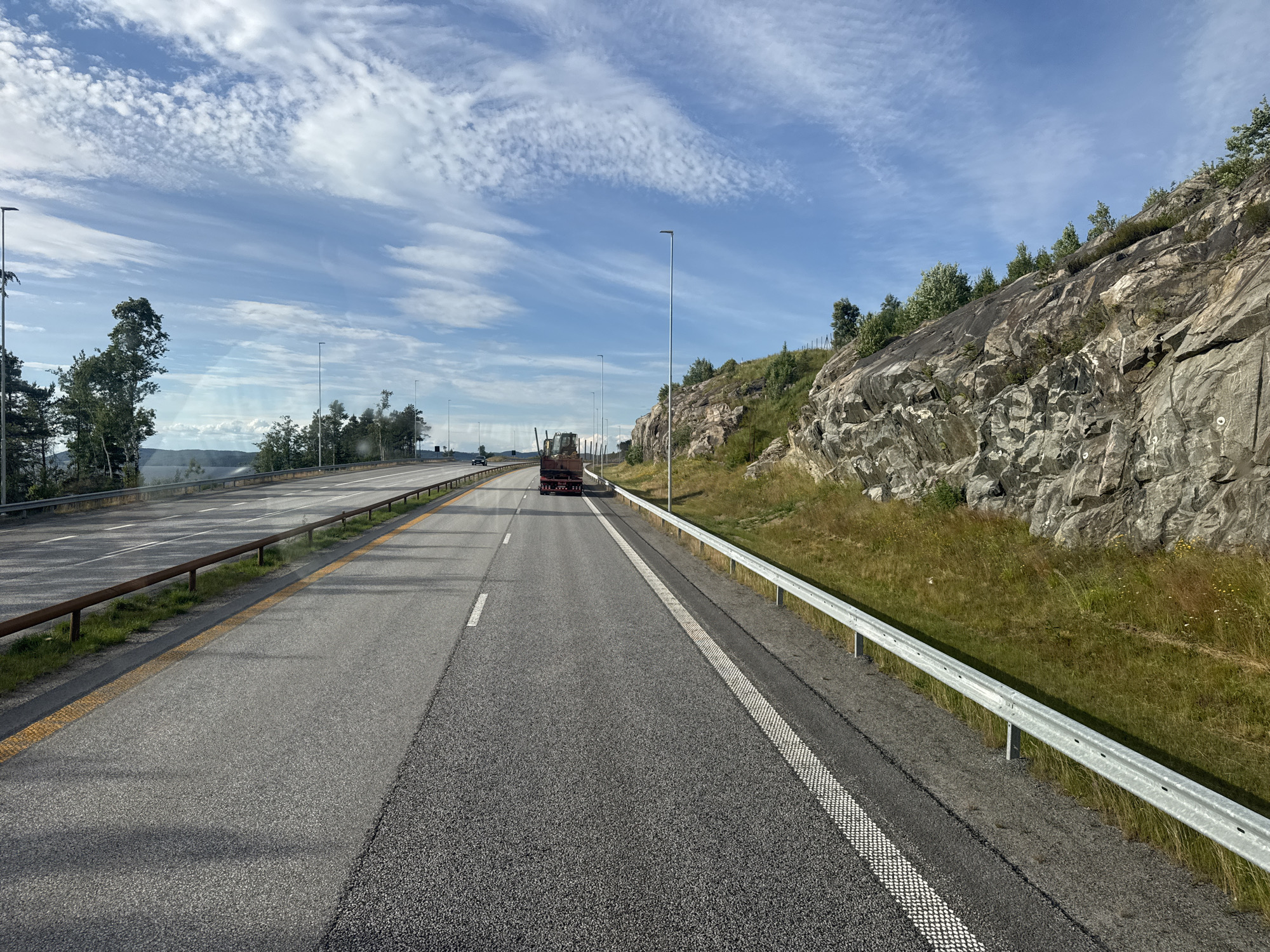 The image shows a scenic view of a highway in Espa, Norway. The highway is paved with asphalt and has multiple lanes, separated by white and yellow lines. A guardrail runs along the right side of the road, and a rocky, tree-covered hillside rises to the right. The sky is blue with scattered white clouds, suggesting it is daytime with fair weather.
In the distance, a red truck with a flatbed trailer is visible driving on the highway. There are no people clearly visible in the image. The overall setting is a highway cutting through a natural landscape, with the rocky hillside and trees indicating a rural or semi-rural location. The time of day appears to be during daylight hours, possibly late morning or afternoon, given the lighting and shadows. The clear sky and bright sunlight suggest pleasant weather conditions. There is no visible text in the image.