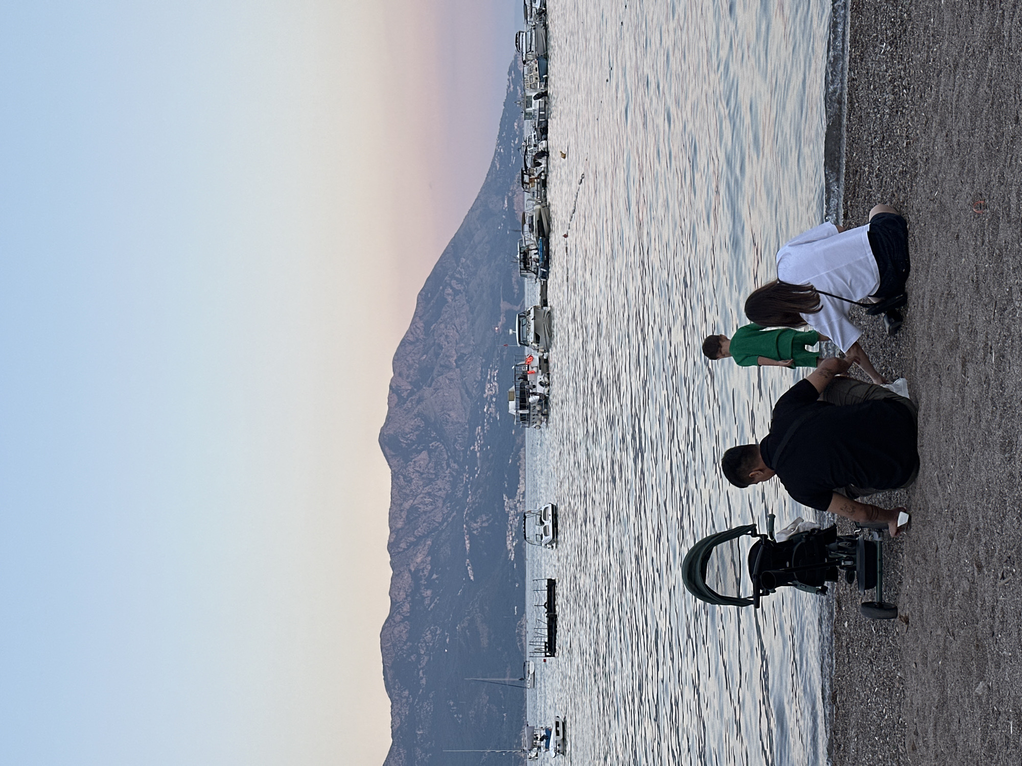 A family is relaxing on a pebble beach in front of the sea in Budva, Montenegro. A father in a black shirt and khaki pants sits with his back to the camera, next to a stroller. A mother in a white shirt and dark shorts sits facing the sea, reaching towards a small child in a green outfit who is standing by the water's edge. In the background, numerous boats are anchored in the calm sea, with a mountain range forming the backdrop. The sky is clear with a gradient of light blue to pale pink.