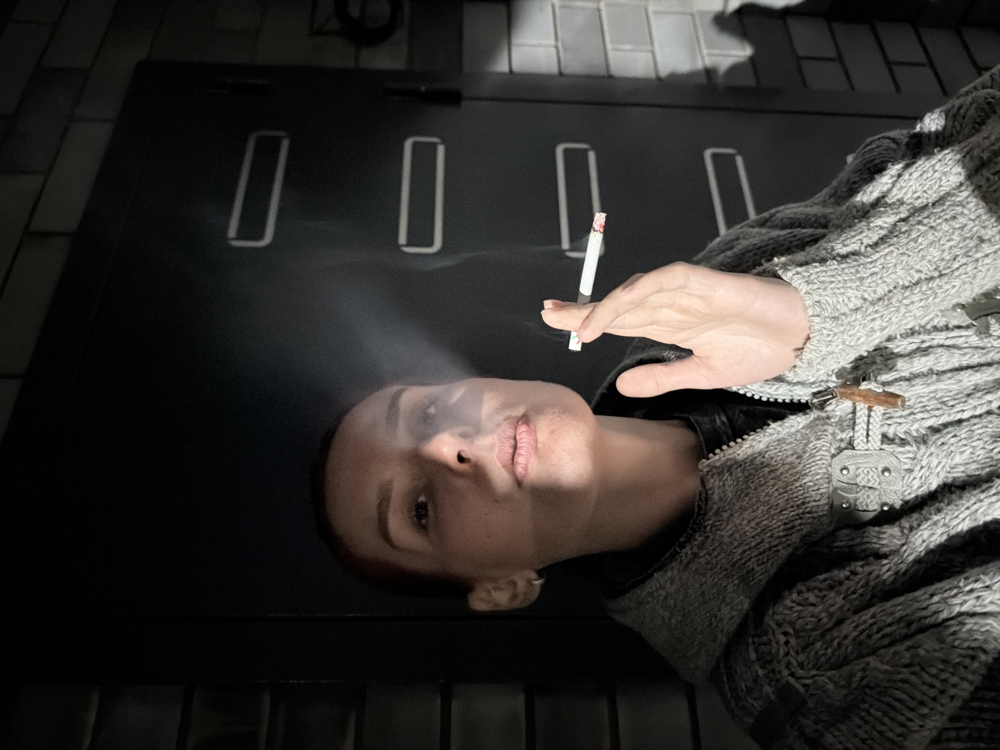 A person, likely a woman with closely cropped hair or a shaved side, is depicted from a top-down perspective, looking directly into the camera. She wears a textured grey knitted jacket or cardigan featuring prominent wooden toggle fasteners. Her hand is visible, holding a lit cigarette from which a plume of smoke rises.
The scene appears to be an intimate indoor setting, possibly a room or a covered area. In the background (or above the subject, given the perspective), a dark, flat surface displays several bright, elongated rectangular outlines, which could be reflective strips or illuminated architectural features on a ceiling or a high wall. A section of light-colored square tiles is also visible, suggesting a tiled ceiling or an adjacent wall.
The person is actively smoking the cigarette. Her expression is neutral and direct, creating a contemplative or self-aware mood. The lighting is dim but dramatic, with a strong light source from above or near the camera illuminating the person's face, the cigarette, and the smoke, casting significant shadows and creating high contrast. The heavy knitwear suggests a cool environment. No visible text is present in the image. The setting is in Chaikovsky, Russia.