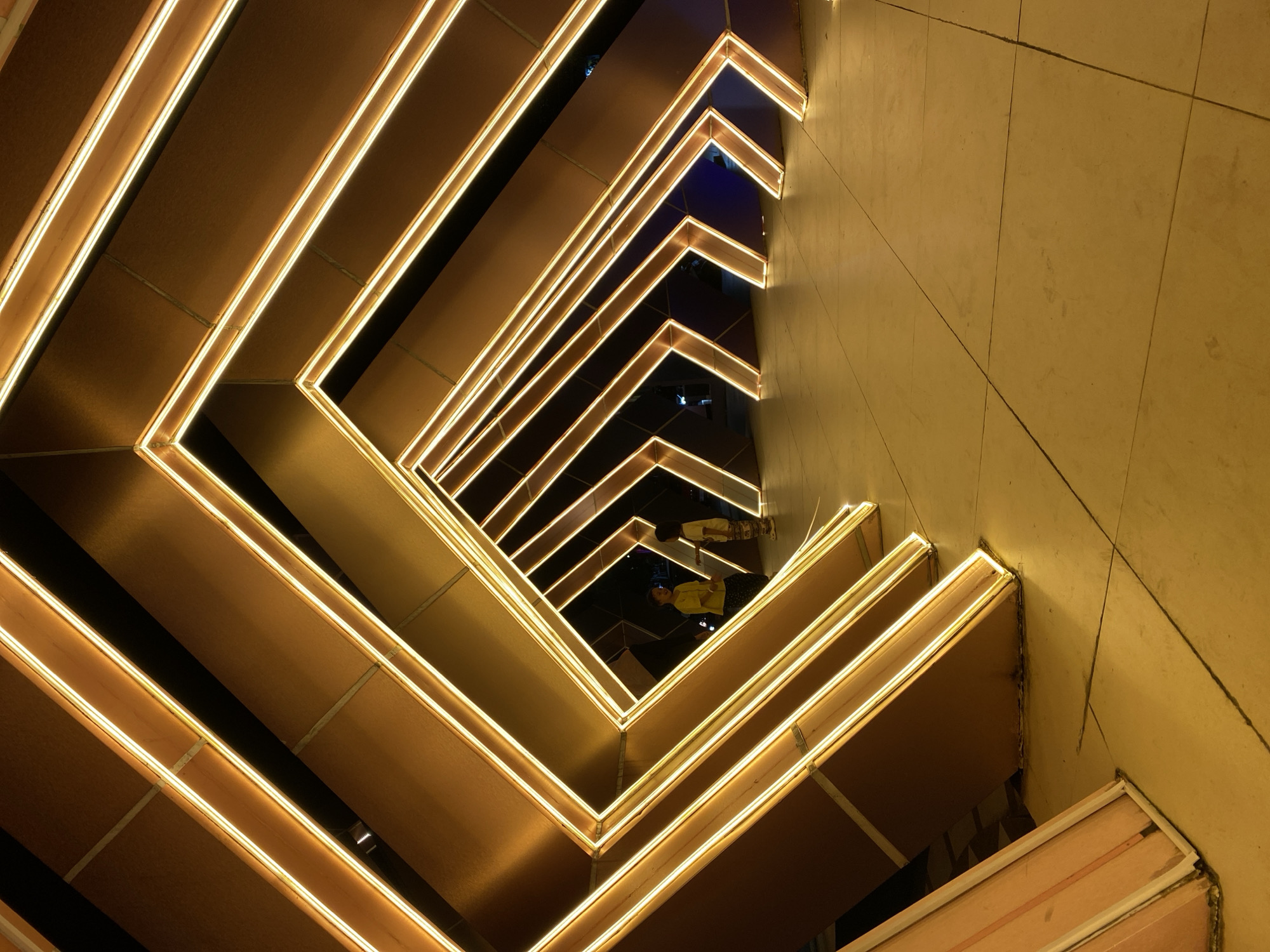 The image captures a modern art installation in Banjarbaru, Indonesia, likely at the Taman Vanide. The installation consists of a series of angled, geometric structures outlined with warm yellow LED lights, creating a tunnel-like effect. The structures appear to be made of a matte, dark material with lighter accents along the edges. The ground is paved with large, light-colored tiles that reflect the warm glow of the lights. Two individuals, possibly children, are visible in the distance within the illuminated tunnel. One is wearing a yellow shirt, and the other is wearing a lighter colored shirt. The overall impression is one of contemporary design and illuminated public art.