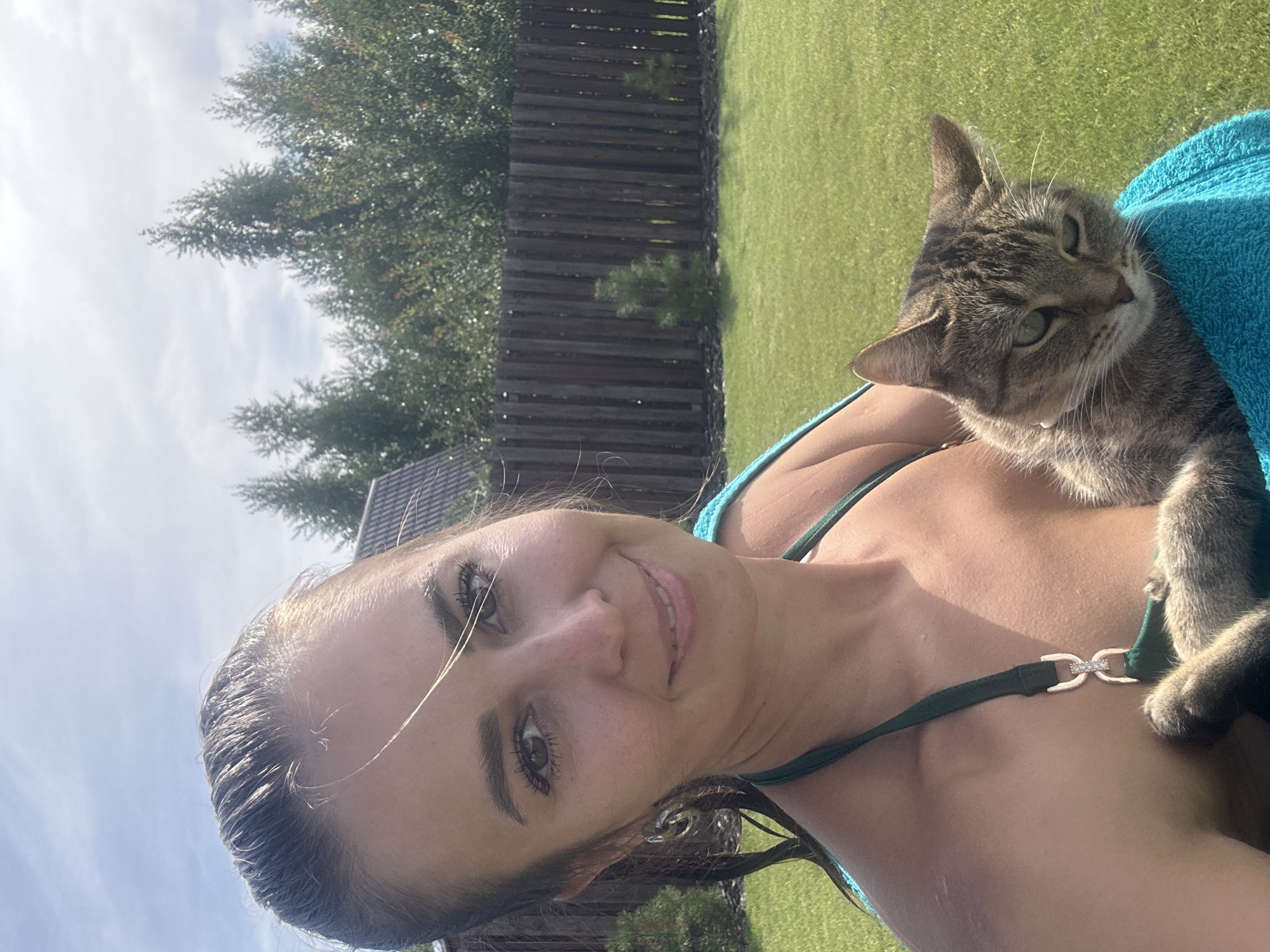 An outdoor, medium shot shows a woman holding a cat while standing in a grassy yard. The woman is in the foreground, facing the camera and smiling. She has long, dark hair that is pulled back, and she is wearing a green bikini. The cat is tabby-colored and is nestled in her arms, wrapped in a blue towel. Behind the woman, a wooden fence runs across the middle ground, with trees and a roofline visible beyond. The sky is partly cloudy, and the sun is shining, casting shadows on the grass.