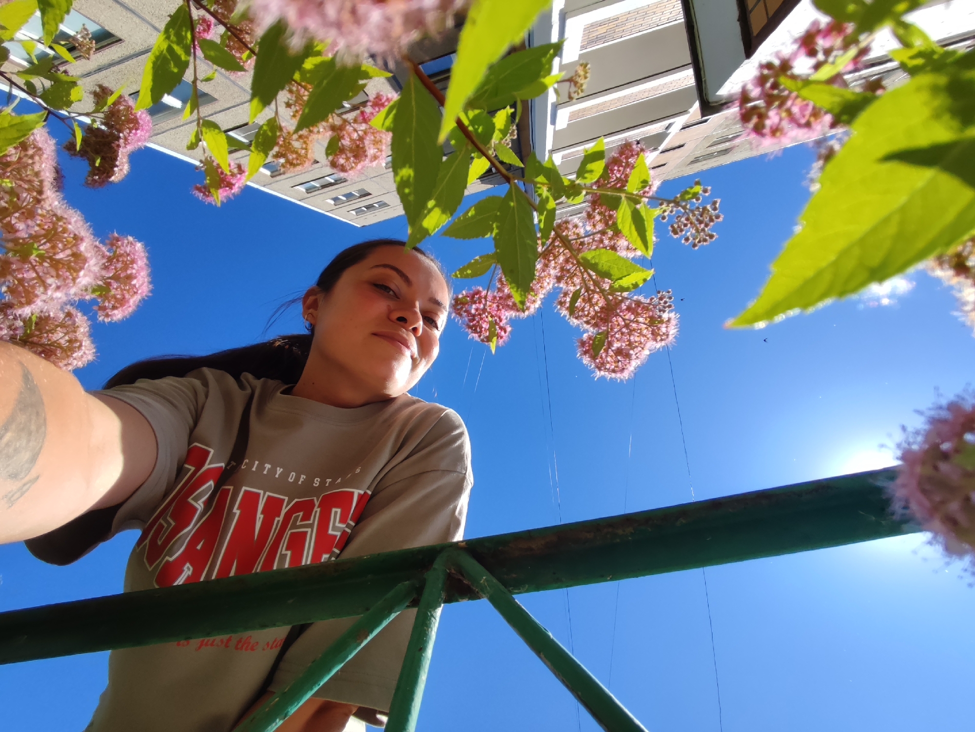 An upside-down, medium shot captures a young woman looking up at the camera, positioned at eye level. She has dark hair and is wearing a light-colored t-shirt with red lettering that appears to say "ANGE." A tattoo is visible on her left arm. The background is a bright blue sky with some wispy clouds and the sun is shining brightly. In the foreground, there is a green railing and branches with small pink flowers and green leaves. The scene is set in front of a building. The orientation of the image suggests it was taken from a low vantage point, possibly looking up from the ground.