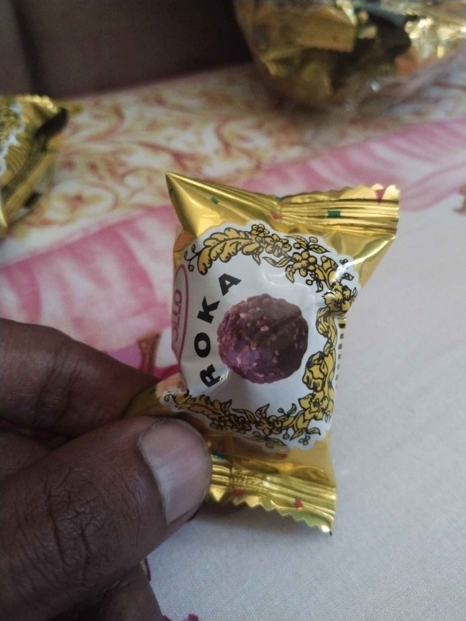 A hand holds a single "OTTO ROKA" chocolate candy wrapped in a gold and white foil wrapper. The candy is dark brown with a textured surface. The background shows more wrapped candies and a patterned surface, possibly a tablecloth. The scene's location is unknown.