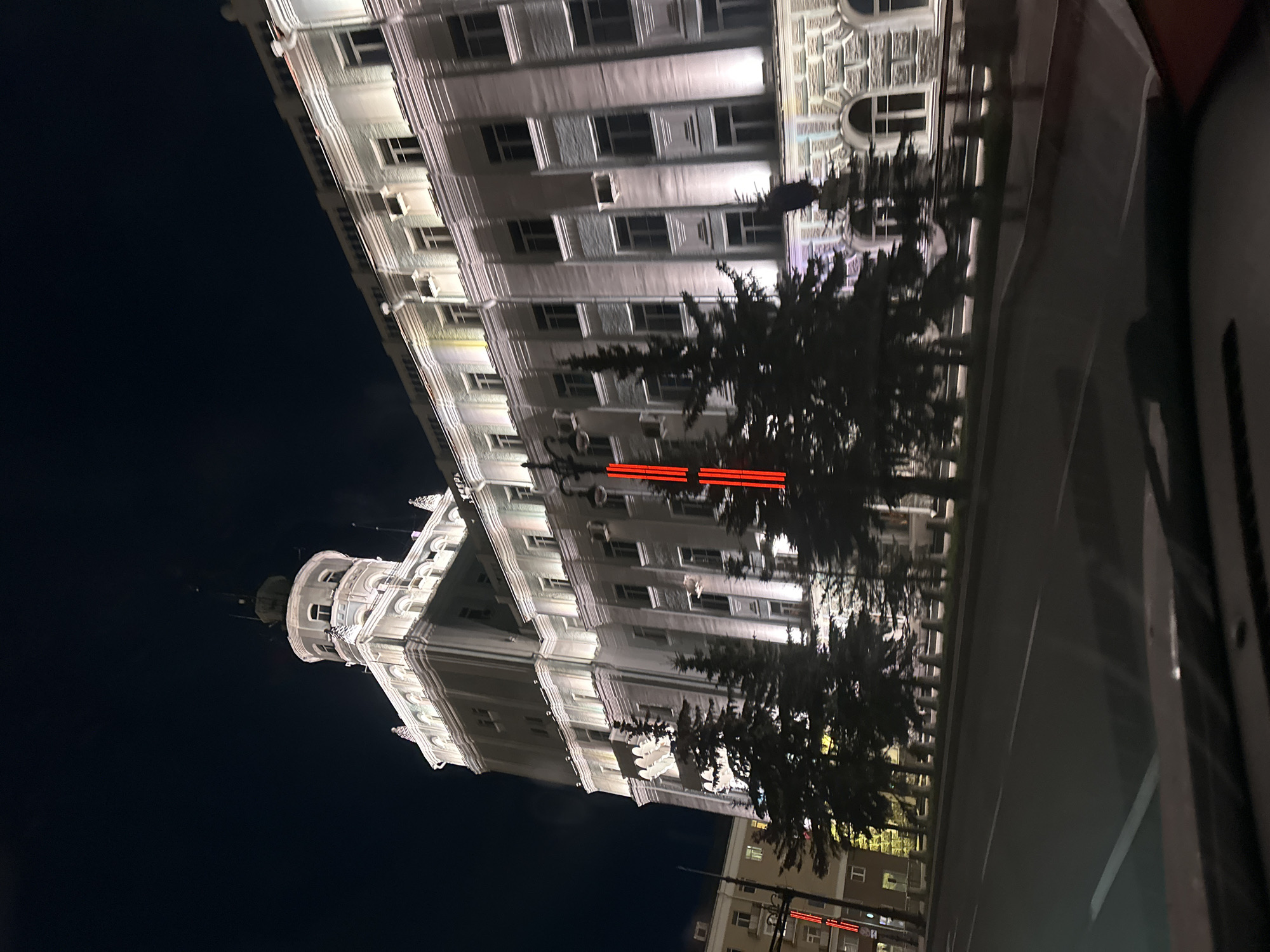 The image shows a nighttime view of the Perm State Art Gallery located in Perm, Russia. The gallery is a large, multi-story building with ornate architectural details and illuminated windows. Trees partially obscure the view of the building, and streetlights are visible. A road runs along the side of the building. Other buildings with illuminated windows are also visible in the distance.