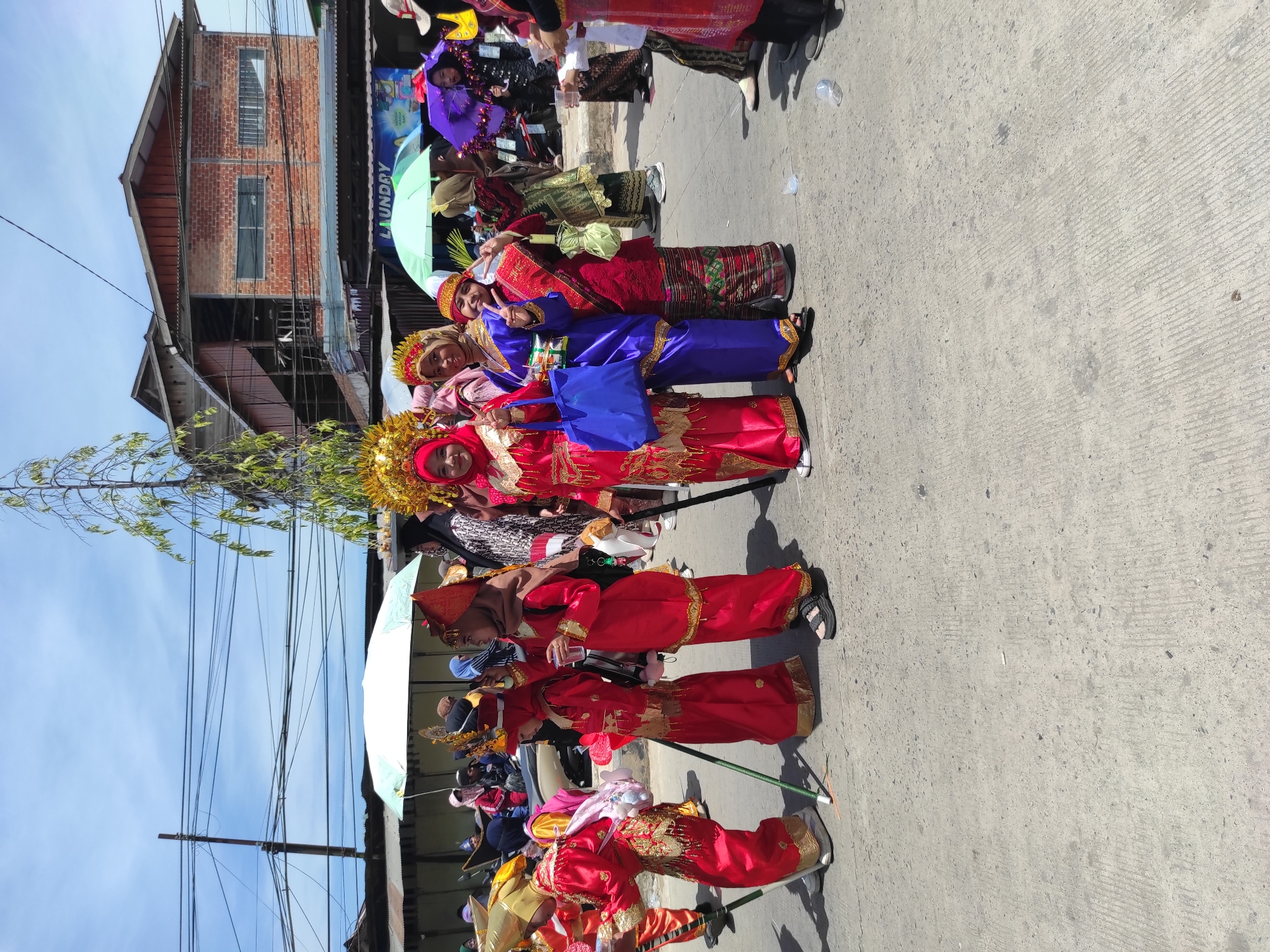 In Muara Jawa Ulu, Indonesia, a group of individuals, primarily young girls, are participating in an outdoor procession. They are dressed in traditional, vibrant red and gold attire, with some wearing elaborate headdresses. Several people are holding long, slender poles. The group is walking on a paved road, with buildings visible in the background, including a brick structure with multiple windows and a sign that reads "LAUNDRY." Several people are standing under umbrellas, suggesting a sunny day. The scene appears to be a cultural celebration or parade.