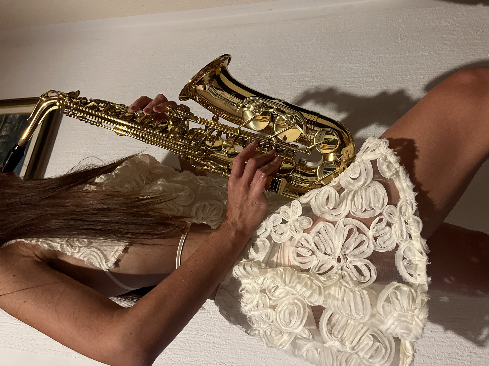A person is holding and playing a saxophone in what appears to be a dimly lit room. The person, whose lower body is visible, is wearing a white, sheer dress with floral embellishments. The saxophone is golden and is positioned in front of the person's body. The background is a textured white wall. There is a framed picture hanging on the wall behind the person. The lighting creates shadows on the wall. The overall scene suggests an artistic or performance setting, potentially indoors. The location in Šušanj, Montenegro is not visually discernible from the provided image.