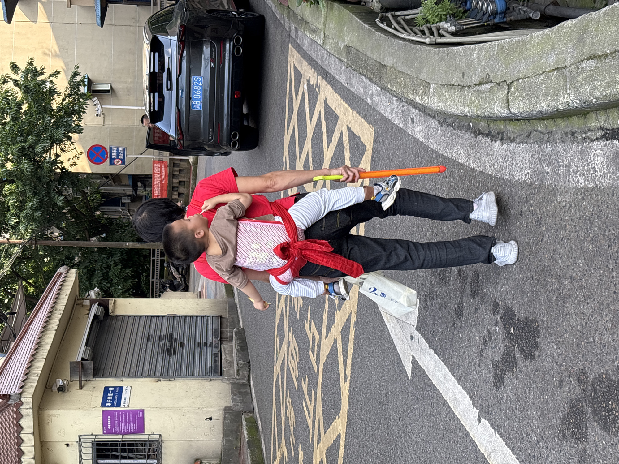In the Yuzhong District of China, a person wearing a red shirt and black jeans walks on a paved street. They are carrying a young child on their back using a red baby carrier. The child is wearing a brown shirt and striped pants. The person is holding a bright orange pole in their right hand. In their left hand, they are carrying a white plastic bag with Chinese writing on it. A dark gray SUV is parked on the right side of the street, and a man is partially visible in the driver's seat. The street has yellow lane markings and a white arrow indicating direction. A building with a brown tiled roof and closed metal shutters is on the left side of the image, with posters on its wall. Trees and other buildings are visible in the background.