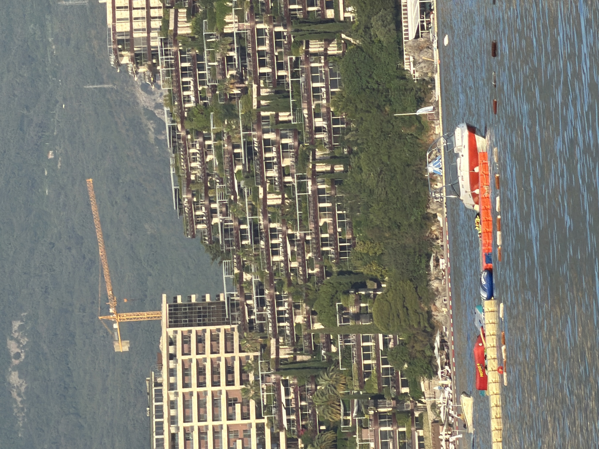 This media file captures a vibrant coastal scene in Budva, Montenegro, under bright, sunny daylight. The image is oriented to show a calm body of water in the lower right, with a sprawling complex of modern buildings and a lush green hillside occupying the left and central parts.
The primary subjects include numerous multi-story buildings, likely residential or resort properties, which feature contemporary architecture with repetitive patterns of balconies and large windows. These structures are integrated into a densely vegetated, mountainous landscape, with trees and shrubs covering the hillside and landscaped greenery, including palm trees, within the complex.
A prominent yellow tower crane stands tall on the left side, situated near one of the buildings, indicating ongoing construction or development in the area.
On the calm water, several boats are visible. A distinctive red and white boat, possibly a yacht or pleasure craft, is moored near the shore. Partially visible text on its side reads "POSEIDON." A series of red and blue inflatable elements, possibly part of a water park or obstacle course, extend across the water, marked by buoys.
The scene suggests a blend of leisure activities, with boats and water features, alongside active development and expansion of the coastal infrastructure. The clear skies and bright illumination indicate pleasant weather conditions.