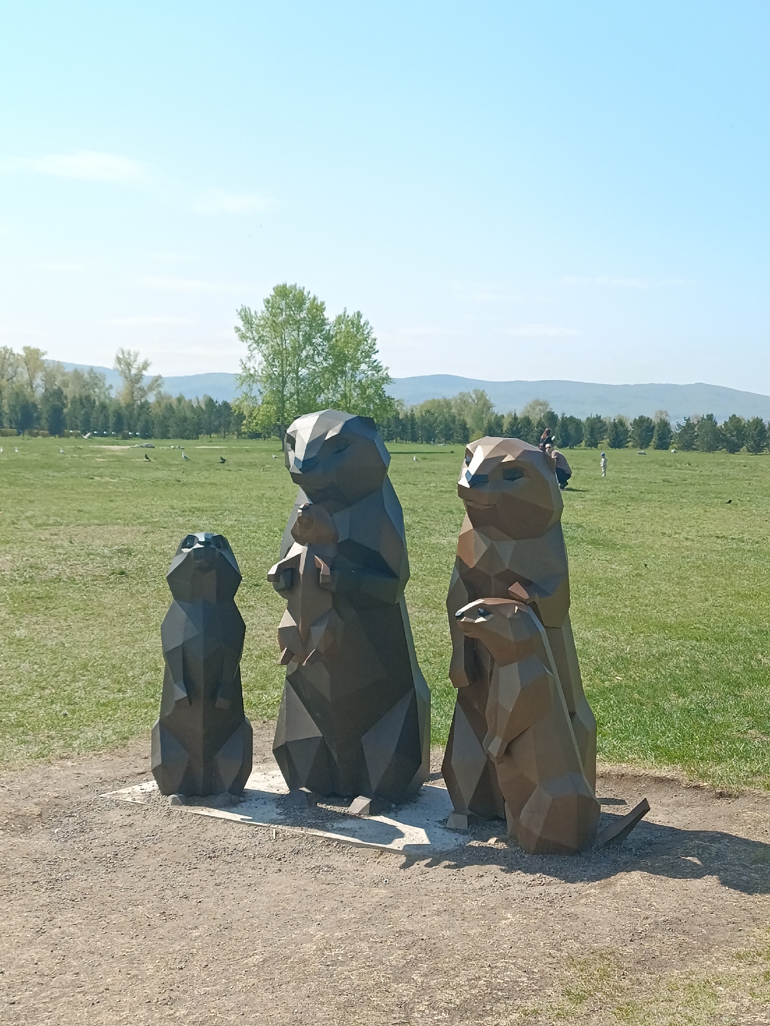 The image shows four geometric metal prairie dog sculptures on a concrete slab surrounded by a gravel path in a grassy field. In the background are scattered trees and distant hills under a blue sky. The sculptures appear to be in a park or open space in Krasnoyarsk, Russia. People are scattered in the background of the grassy field. The statues are varying shades of brown.