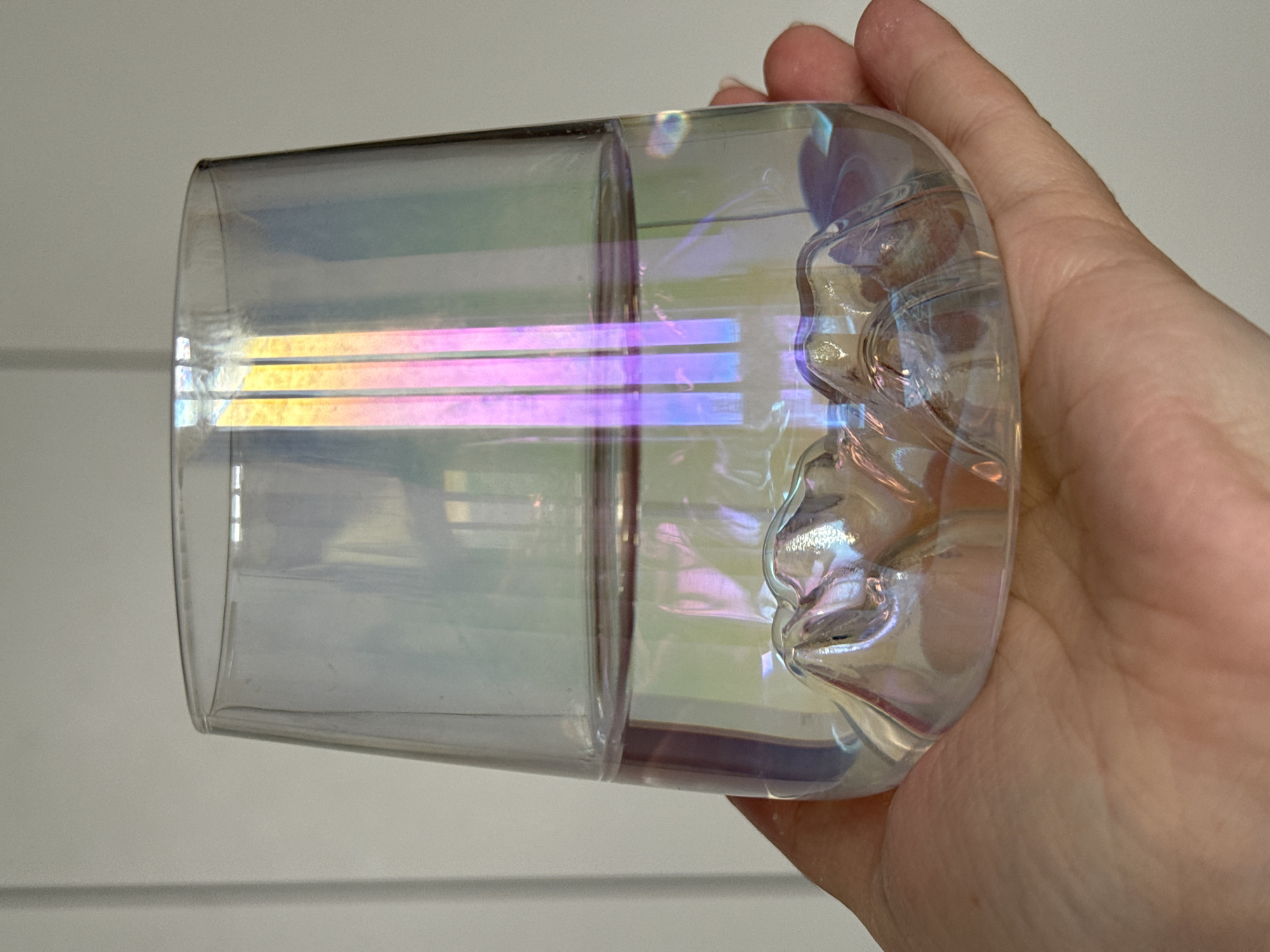 A hand holds a clear, iridescent glass or cup horizontally against a plain, light-colored background that shows faint horizontal lines. The object exhibits a strong iridescent effect, displaying a spectrum of colors including yellow, pink, and purple, particularly visible in distinct horizontal bands on the left side of the glass. The right side of the glass features a textured, sculpted, or wavy surface, which reflects light in a more fragmented and complex manner. The hand, likely a right hand, has light skin tone with visible skin texture and creases. The fingernails are short and unpolished. The lighting appears to be indoor, enhancing the colorful reflections and refractions within the glass. There are internal reflections, possibly of light sources, visible as bright vertical and horizontal lines within the glass. The glass construction suggests it might be double-walled or have a discernible internal structure, as indicated by a vertical line or seam in the middle. No specific activities other than holding the glass are apparent. There is no visible text.