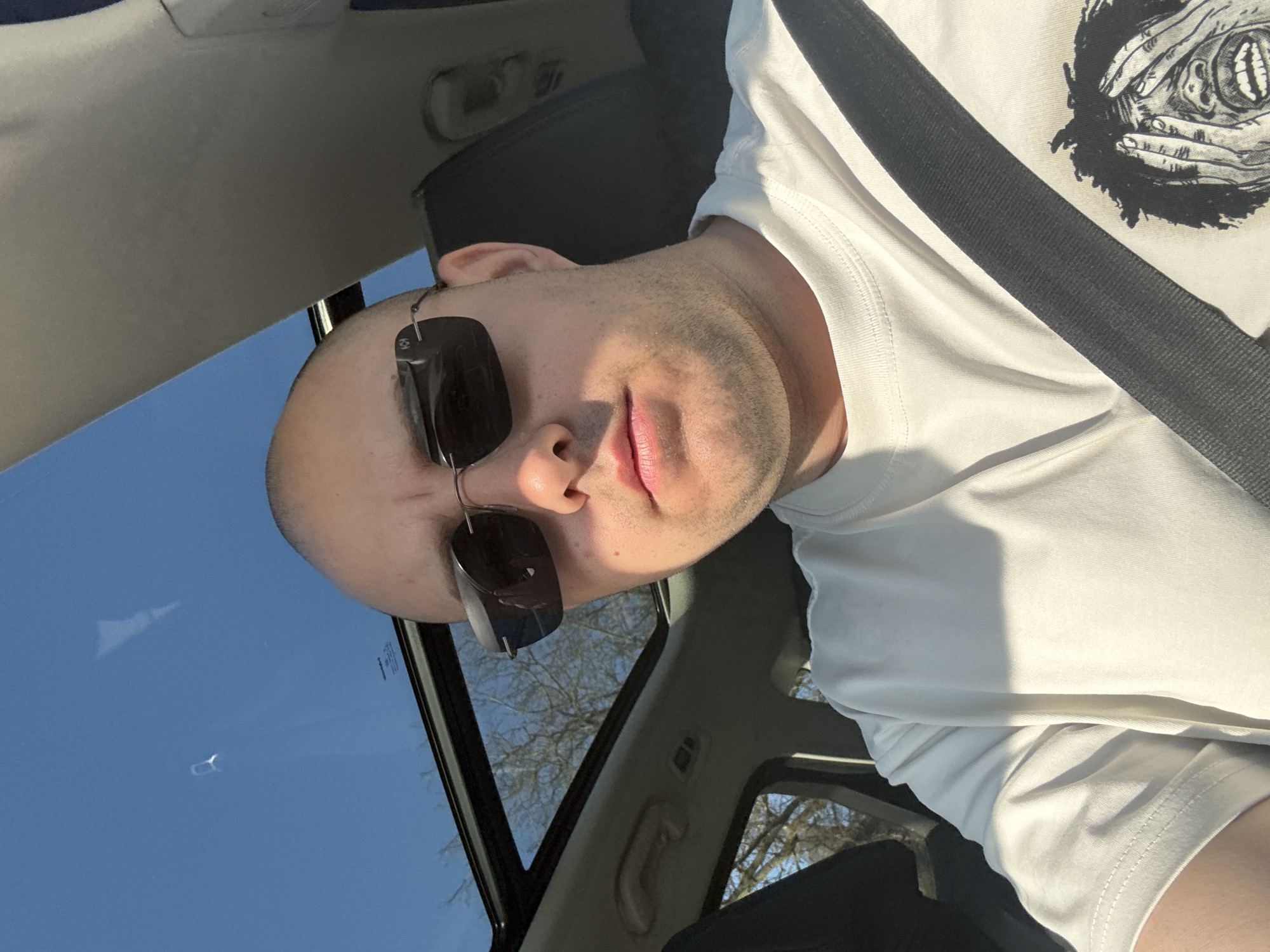 A bald man with sunglasses and a white t-shirt with a black graphic print is in a car with a black seatbelt fastened. The sky is visible through the sunroof. The image does not provide information to determine the location or any information about the city of Pleven, Bulgaria.