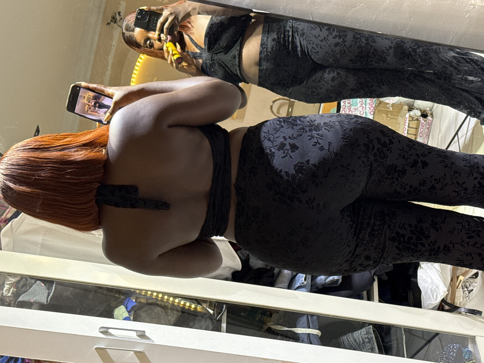 The image shows a woman in a black floral-patterned outfit standing in a room. She is holding a smartphone in her right hand and taking a picture of herself in a mirror. Her reflection is visible in the mirror, which also shows a yellow object, possibly a makeup brush, in her hand. The room appears to be a bedroom or dressing area, with a wardrobe on the left side of the frame containing various clothing items. The overall lighting suggests an indoor setting. The city of Abuja, Nigeria is not explicitly depicted or suggested in the image.