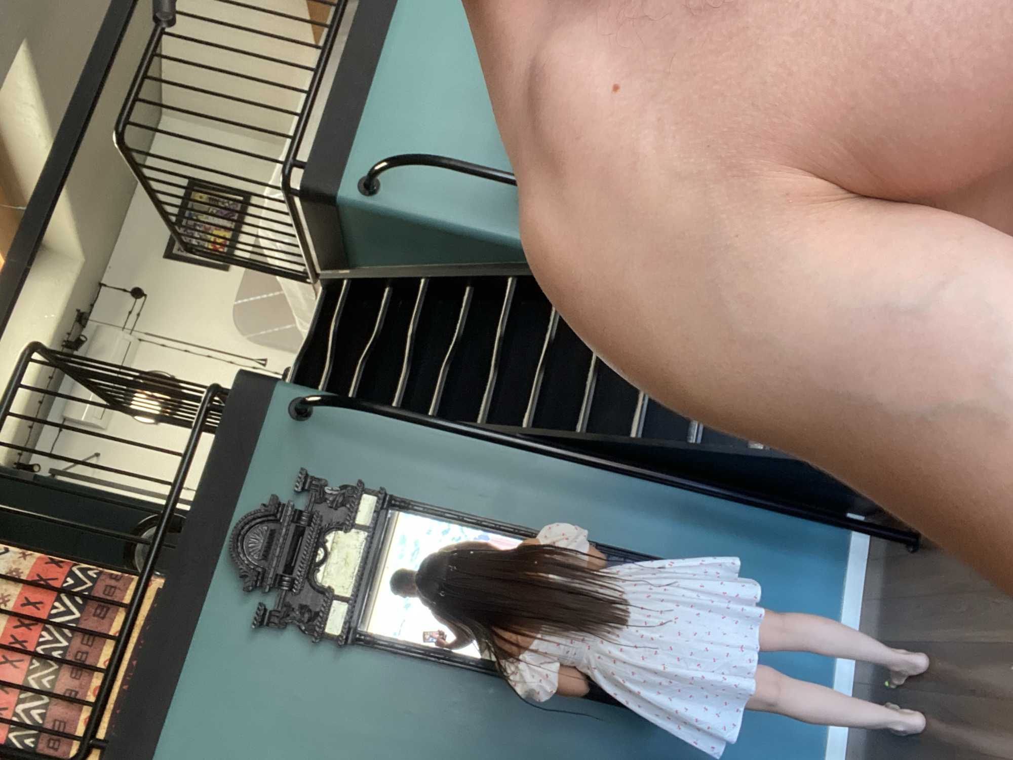 The image shows a person holding a phone and taking a photo of themselves in a mirror. The person is wearing a white dress with red polka dots. They have long, dark hair. In the background, there is a staircase with black railings. The walls are painted a teal color. There is a framed picture on the wall above the staircase. The floor is made of wood. The overall scene appears to be indoors, possibly in a home or a public building. The lighting suggests it is daytime. There is no visible text in the image.
