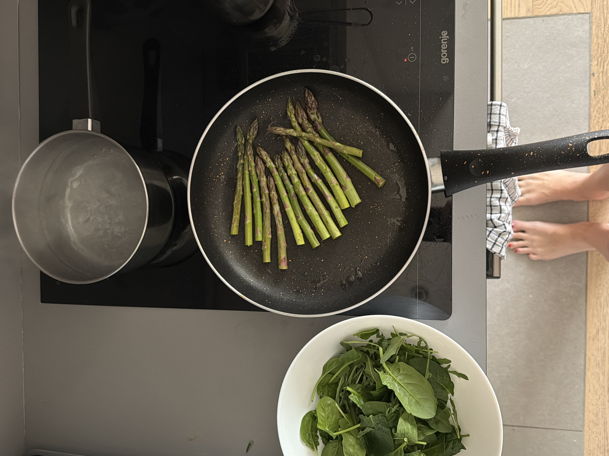 An overhead shot captures a cooking scene in a modern kitchen, situated in Belgrade, Serbia. The primary elements are centered around a black induction stovetop, visibly branded "Gorenje" and displaying a small "i°" symbol. On the left burner, a silver saucepan contains water, suggesting it's either heating or has recently been used for boiling. On the right burner, a black non-stick frying pan holds a pile of fresh green asparagus spears, some showing signs of light cooking or seasoning. The pan's black handle, speckled with lighter dots, extends towards the right.
Below the stovetop, on a grey countertop, a white bowl is filled with vibrant green leafy vegetables, appearing to be a mix of spinach and potentially arugula, indicating the preparation of a salad or another fresh component of a meal. A white and blue checkered kitchen towel is draped over the edge of the stovetop unit on the right.
To the far right, a pair of bare feet with red-painted toenails are visible on the kitchen floor, suggesting a person is actively engaged in the cooking process. The floor transitions from a light grey tile directly in front of the stovetop to a lighter wooden floor further into the room. The image conveys a calm, functional moment of preparing a healthy meal.