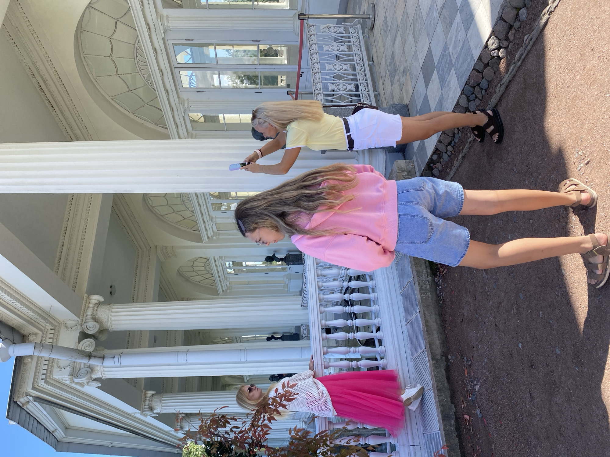 The image depicts three individuals in front of a grand white building with classical architecture, characterized by large columns and ornate detailing. A woman in a white crocheted top and a vibrant pink skirt stands on a railing, looking to her left. To her right, another woman, facing away from the camera, wears a pink hoodie and denim shorts, with her back to the viewer. A third woman, positioned to the right and facing the viewer, is taking a photograph with her phone, wearing a yellow t-shirt and white shorts. The setting appears to be an outdoor area with stone paving and some gravelly ground. The architecture suggests a historical or cultural landmark, potentially within the city of Pushkin, Russia, known for its numerous palaces and parks.