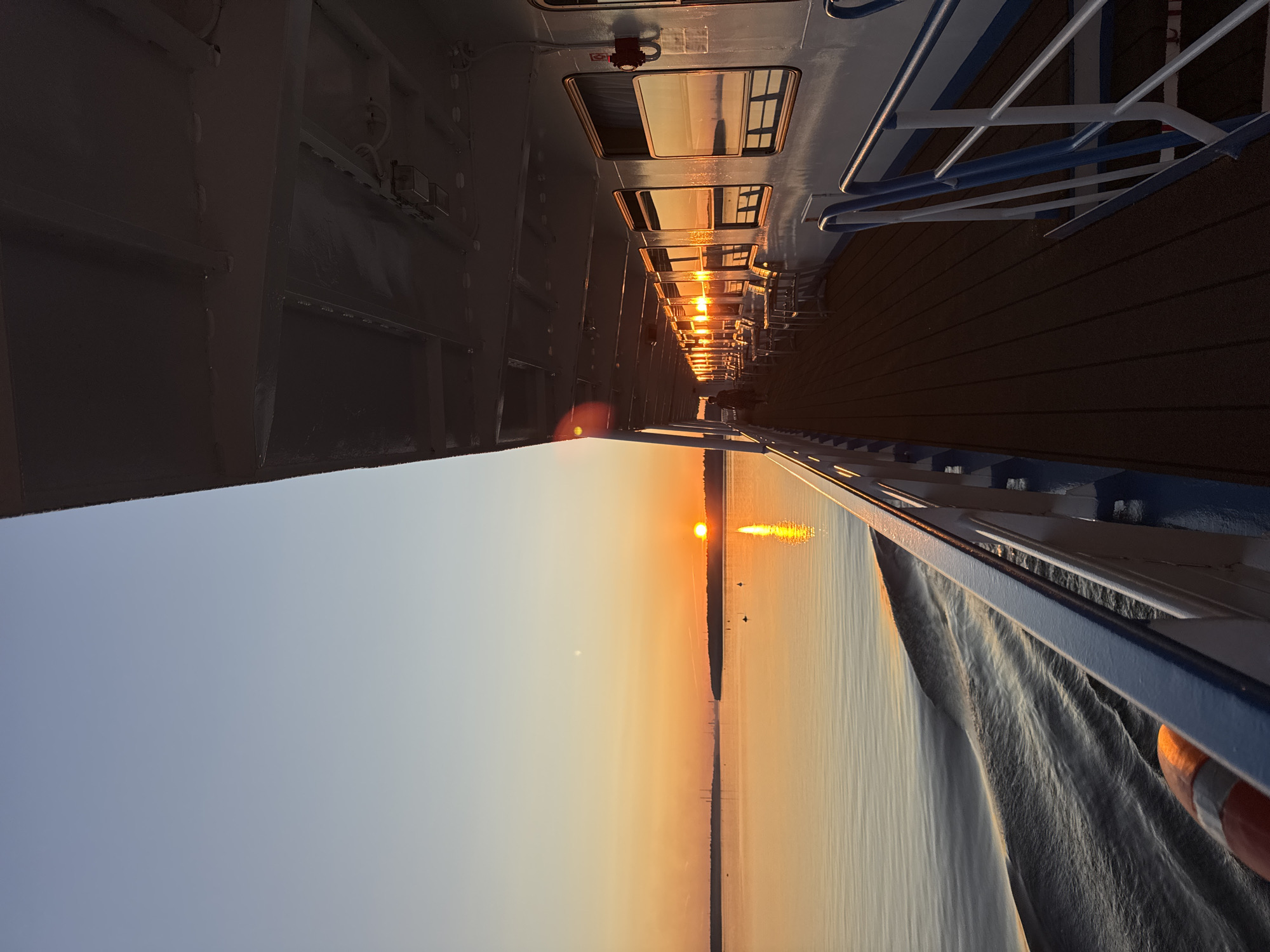 The image depicts a view from the deck of a ship sailing on a body of water at sunset. The sun, a bright orange orb, is low on the horizon, casting a warm glow across the sky and water. The sky transitions from a pale blue at the top to a vibrant orange near the horizon. Silhouetted against the sky are distant landforms, appearing as dark, flat shapes.
The right side of the image shows the side of the ship, with a long row of windows reflecting the sunset. Tables and chairs are visible on the deck in front of the windows. A walkway with railings runs along the side of the ship. The deck itself is wooden.
In the water, a wake trails behind the ship, creating ripples and patterns on the surface. A life preserver is visible near the railing in the foreground.
There is one person visible walking along the deck in the distance. The scene appears to be serene and peaceful. There is no information provided within the image itself to indicate a specific location within the city of Шайма, Russia.