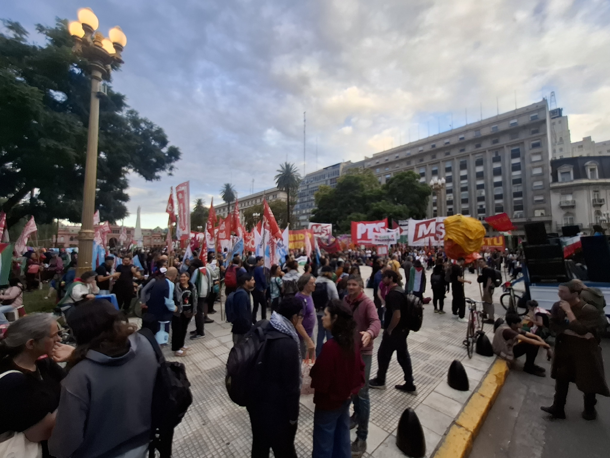 The image depicts a daytime outdoor scene in Buenos Aires, Argentina, at the Plaza de Mayo. Numerous people are gathered, suggesting a protest or rally. Many are holding red flags and banners, some with white lettering. A yellow inflated object is visible amidst the crowd. The background includes the Casa Rosada presidential palace and other large buildings. A tall street lamp with illuminated globes stands to the left. People are standing, walking, and some are sitting on the ground. The weather appears overcast.