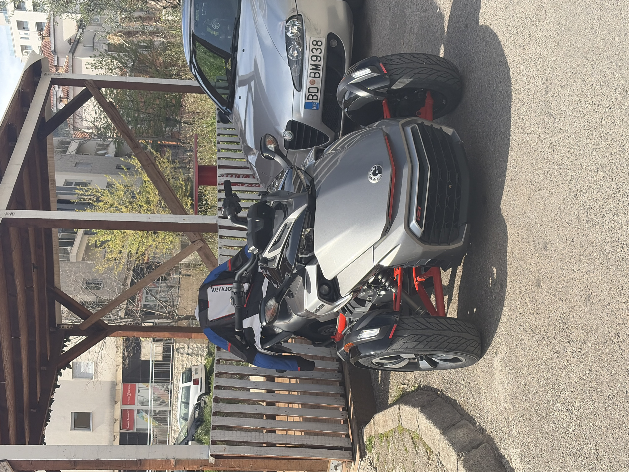 The image depicts a gray Can-Am Spyder parked beside a silver car with license plate BD BM 938. The vehicles are situated near a wooden structure, possibly a covered parking area, and a white fence. In the background, buildings and trees are visible, suggesting an urban or suburban setting in the city of Unknown, Unknown. The lighting indicates it is daytime. A jacket is draped over the handlebars of the Spyder. The image is taken from a high-angle perspective and rotated, showing the Spyder and car partially obscured by the wooden structure.
