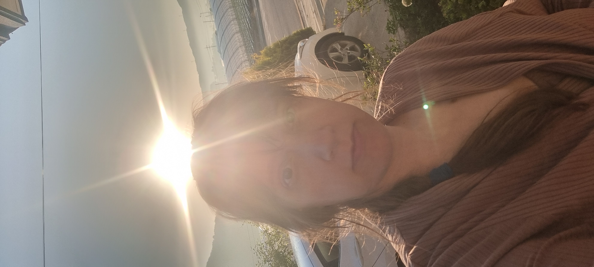 A person is visible in a sunlit, outdoor selfie. The sun's glare obscures details of their face. They are wearing a brown, textured garment. In the background, there is a landscape with a white car partially visible, along with some greenery and a wire-frame structure. The location is potentially Dunnae-myeon, South Korea, based on available information.