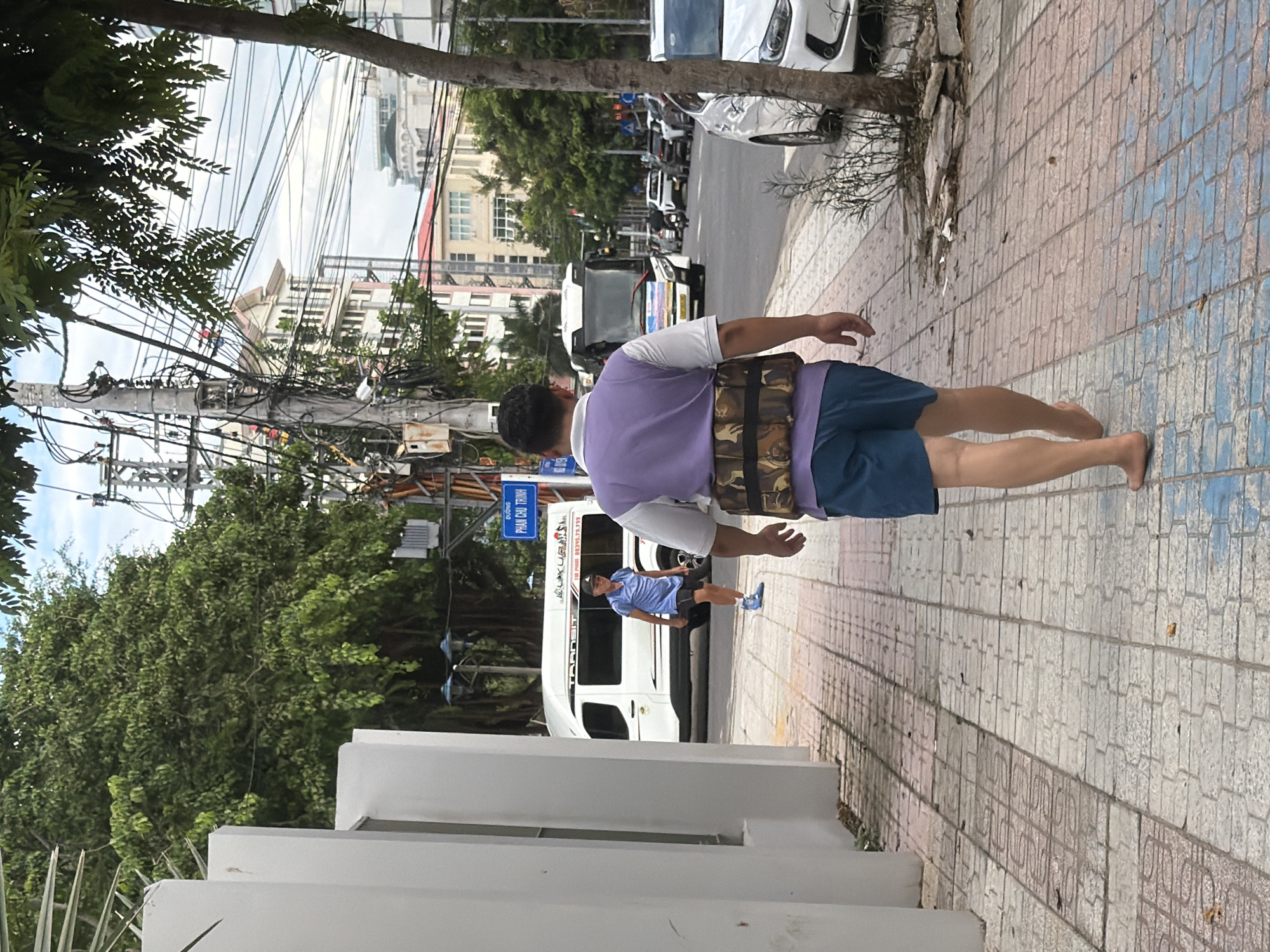 In Nha Trang, Vietnam, a man walks barefoot on a sidewalk. He is wearing a purple vest over a white shirt and blue shorts. A weighted belt is around his waist. In the background, a white van with the word "TRANSIT" on its side is parked. Another man in a blue shirt and black shorts is standing near the van. The sidewalk is made of patterned tiles. The scene is on a street with trees, utility poles, and buildings.