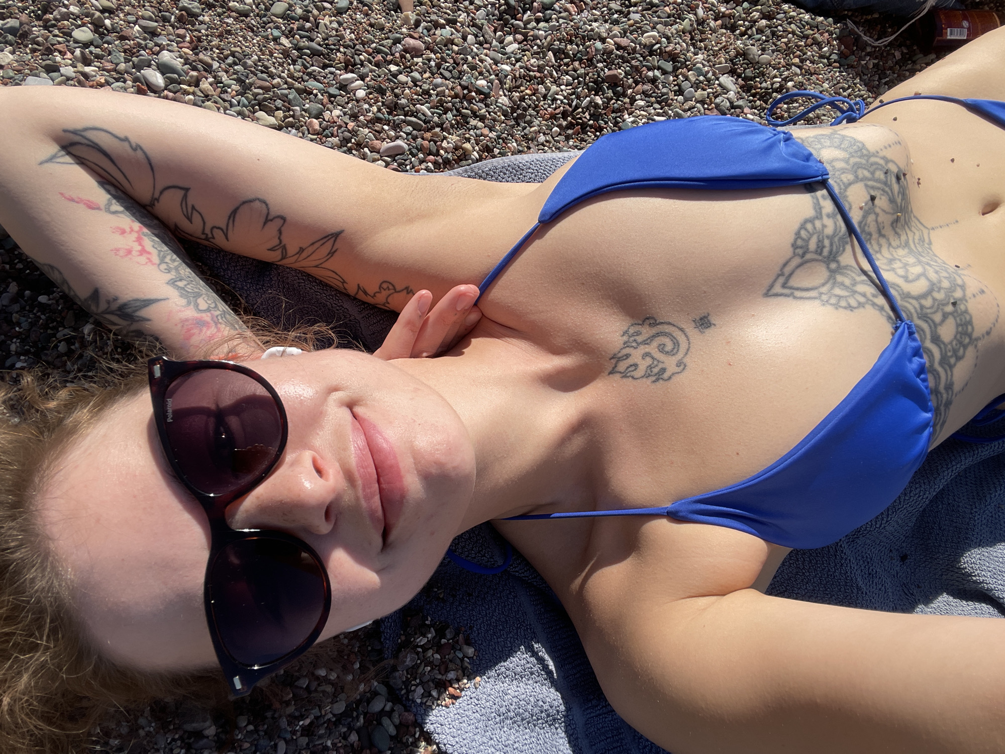 A person is depicted from a high-angle perspective, lying on their back on a pebble beach under bright sunshine, likely sunbathing or relaxing. The individual, who appears female, wears a bright blue string bikini top. Their light-colored, curly hair is spread around their head, and they have dark sunglasses with the brand "Polaroid" visible on the frame. A faint, relaxed smile is present on their lips.
Numerous tattoos are visible across the subject's body. Both arms feature intricate floral or leafy designs. A large, ornate, mandala-like tattoo covers a significant portion of the right side of the torso and rib cage. A smaller tattoo, possibly a stylized symbol or character, is located on the upper chest. A small white earbud is visible near the left ear. The person rests on a light blue or gray textured towel.
The background consists of a dense scattering of small, smooth pebbles and stones in various natural shades, typical of a coastal setting. The strong highlights and shadows indicate clear, sunny weather during daytime. The scene evokes a sense of leisure and tranquility at a beach, specifically in Rafailovići, Montenegro.