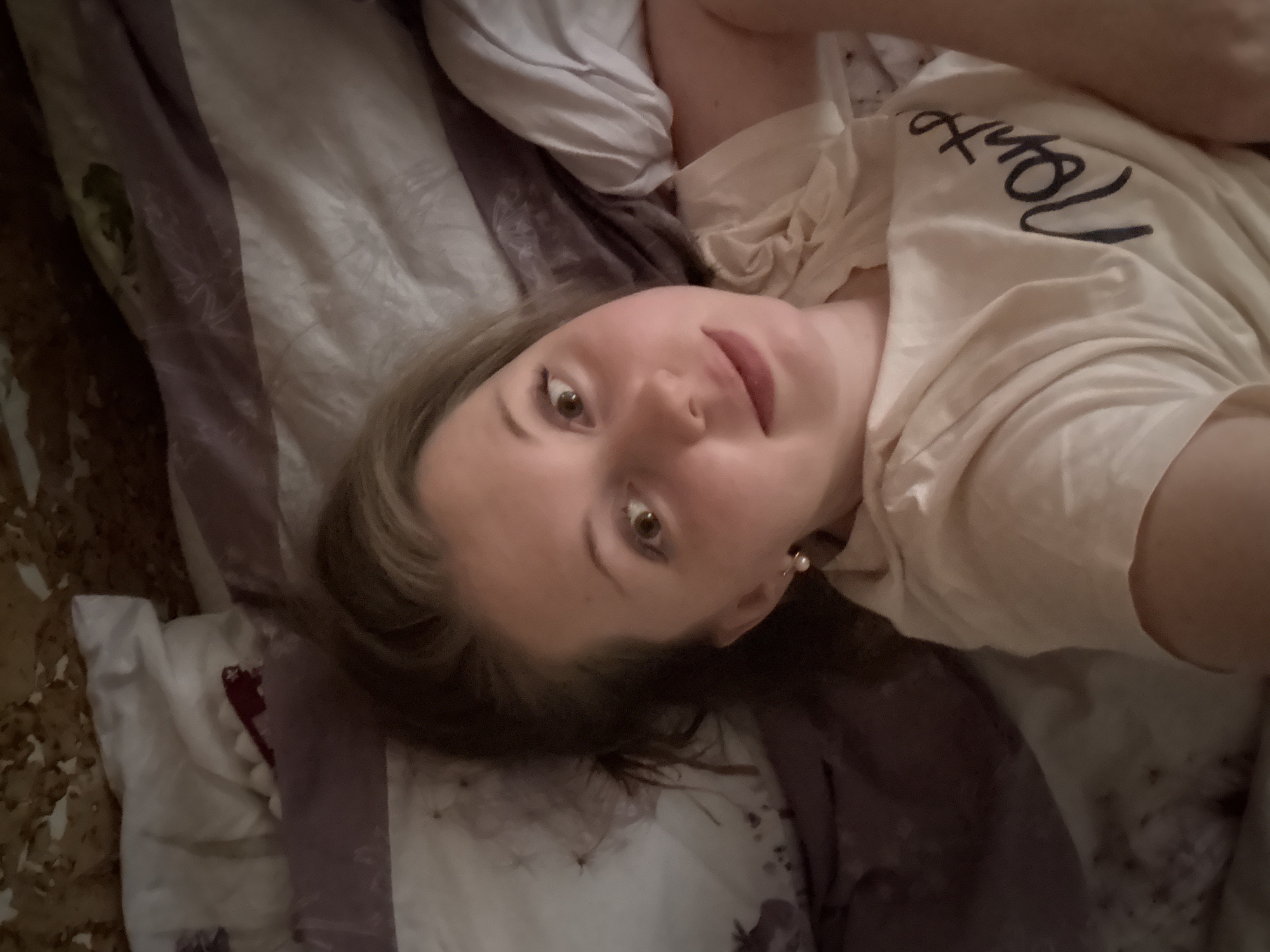 A casual, overhead shot depicts a young woman lying down, possibly in bed. She has light brown hair and is looking directly at the camera with wide, dark brown eyes and a neutral expression that hints at a slight smile. She wears a light-colored (cream or pale yellow) t-shirt, which features the word "Hello" in dark, script-like text across the chest, appearing partially reversed due to the angle. A single pearl earring is visible in her right ear.
The setting appears to be indoors, likely a bedroom, characterized by the bedding beneath and around her. The sheets display a pattern of wide stripes in shades of purple and white, alongside sections with darker, intricate designs in browns and greens. The lighting is soft and subdued, suggesting an indoor environment without harsh direct light. The overall mood is relaxed and informal, typical of a personal photo or selfie taken while resting. The location is unknown.