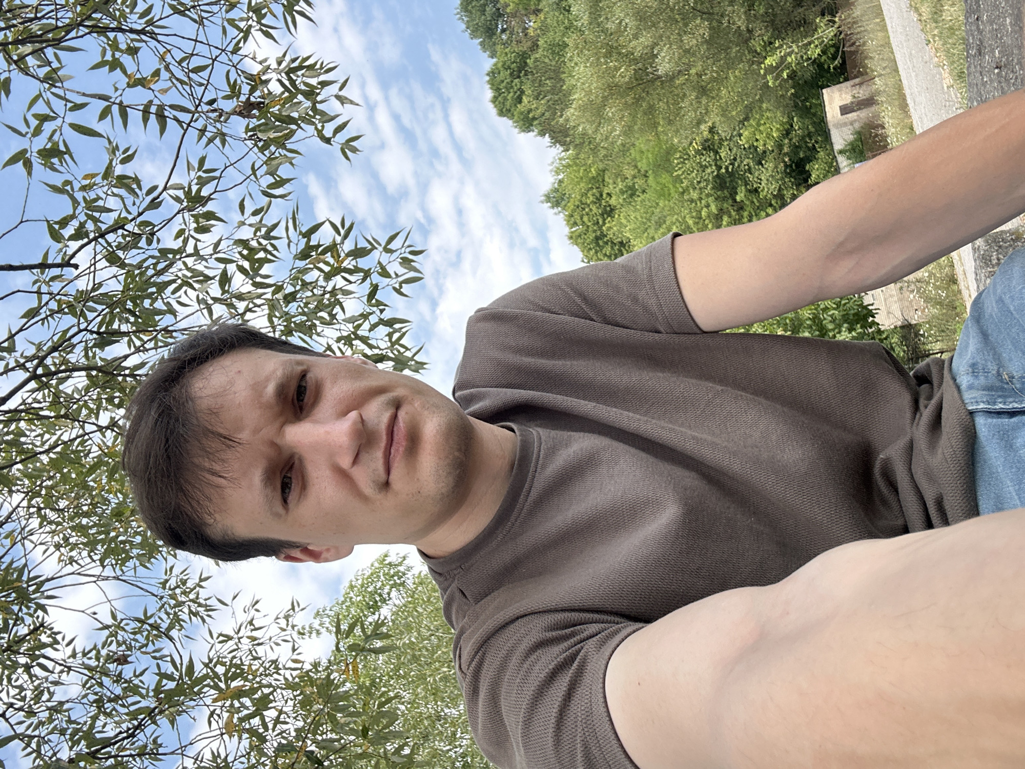 A medium shot captures a man from the chest up, outdoors, with his right arm extended towards the camera, holding it. He is looking directly at the viewer with a neutral expression. He has short, dark brown hair and is wearing a dark brown, textured t-shirt and blue jeans. Behind him, the scene transitions from the dappled sunlight filtering through green leaves and branches of a tree overhead, to a bright, cloudy blue sky. Further in the background, a dense forest of green trees is visible, with a small brick building with a doorway and a fence line in the distance. The overall setting appears to be a park or natural area, possibly on the outskirts of a town. The location is identified as Makiv, Ukraine.